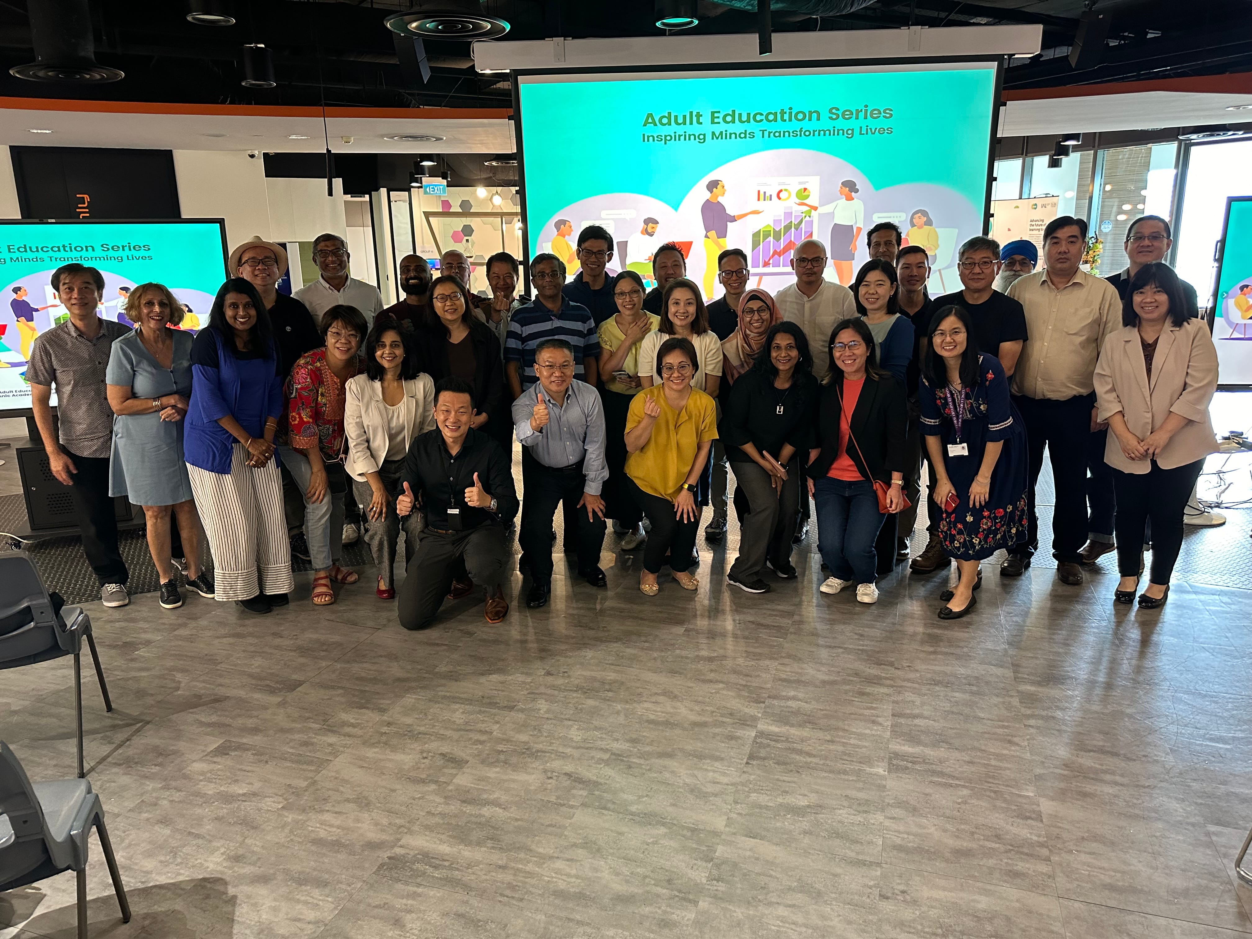A large group photo of approximately 30 participants in a modern event space. They are standing in front of a large digital screen that displays the title 'Adult Education Series: Inspiring Minds Transforming Lives'. Several participants are kneeling or gesturing with thumbs-up and heart signs