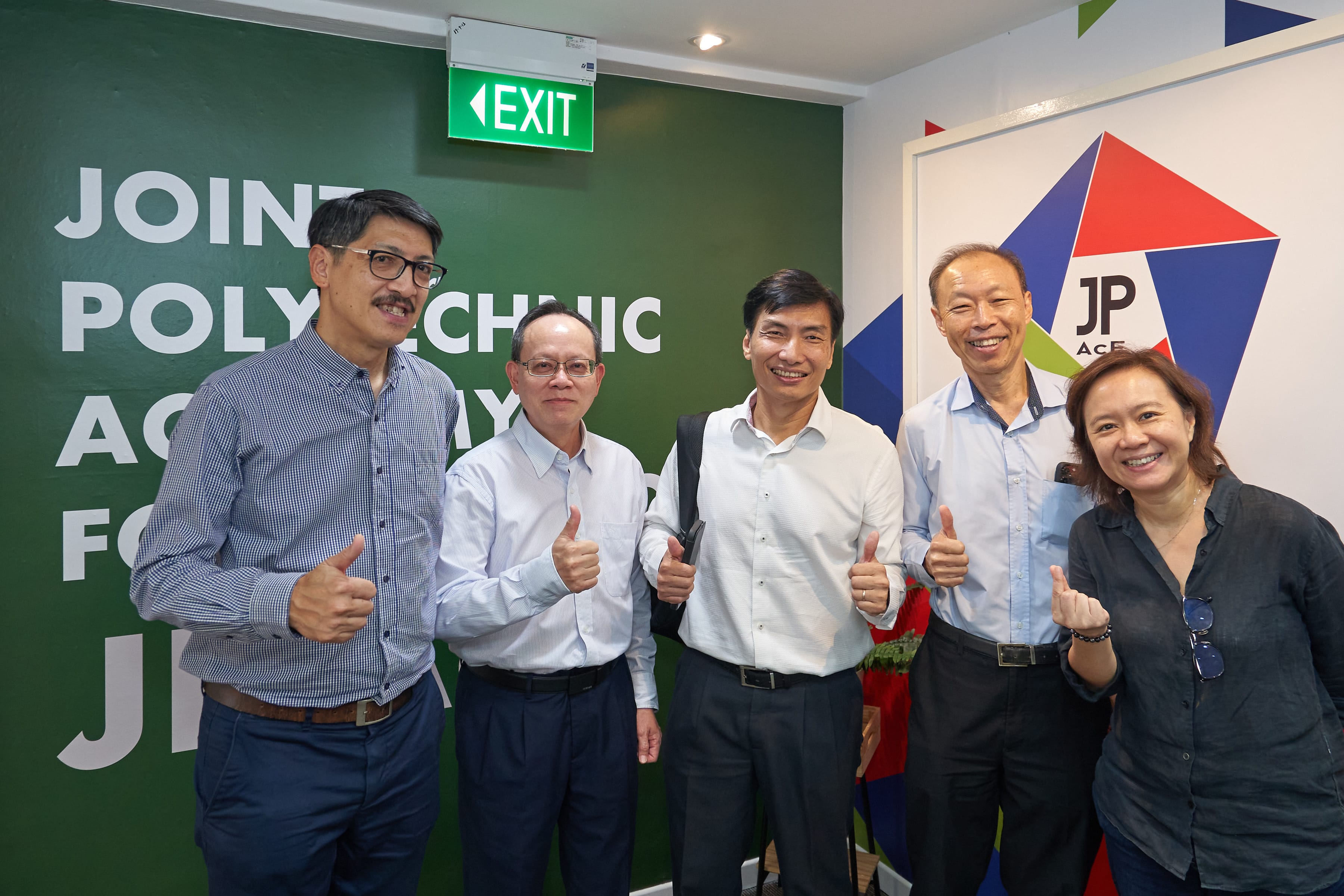 A group of five people, four men and one woman, pose together, all smiling and giving thumbs-up gestures. They are standing against a background that features a prominent green wall with white lettering and a 'JP-AcE' logo with colorful triangles, matching the branding seen in similar event photos.