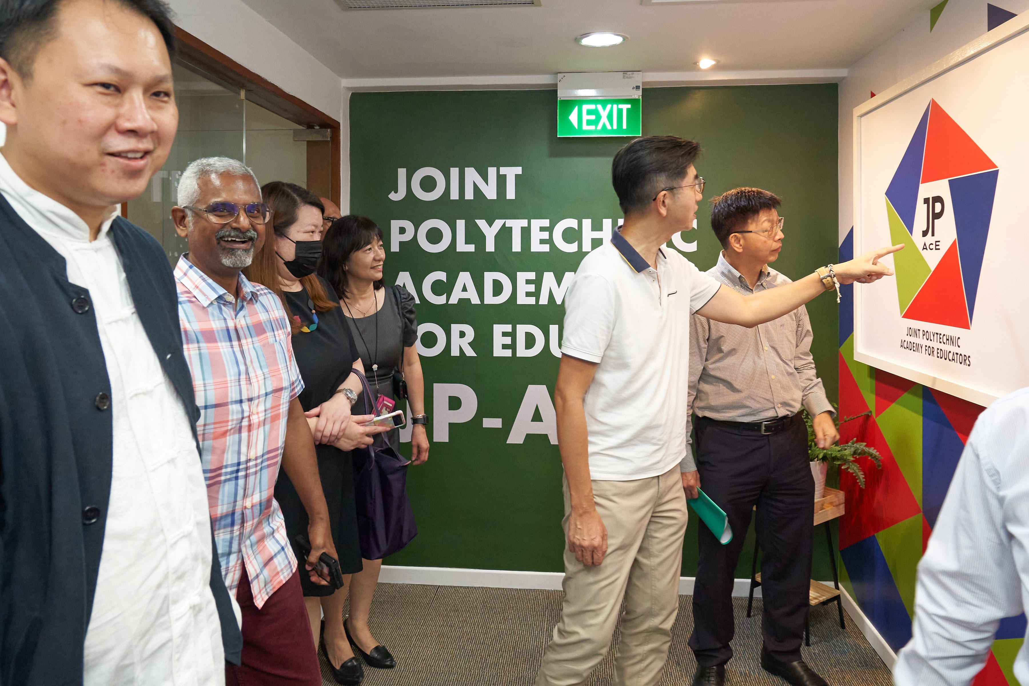 A group of six people stand near an office entrance. A green wall is painted with large white letters reading 'JOINT POLYTECHNIC ACADEMY FOR EDUCATORS' and 'JP-A'. One man in a white shirt points at a colorful pentagon-shaped 'JP-AcE' logo mounted on the wall to the right.