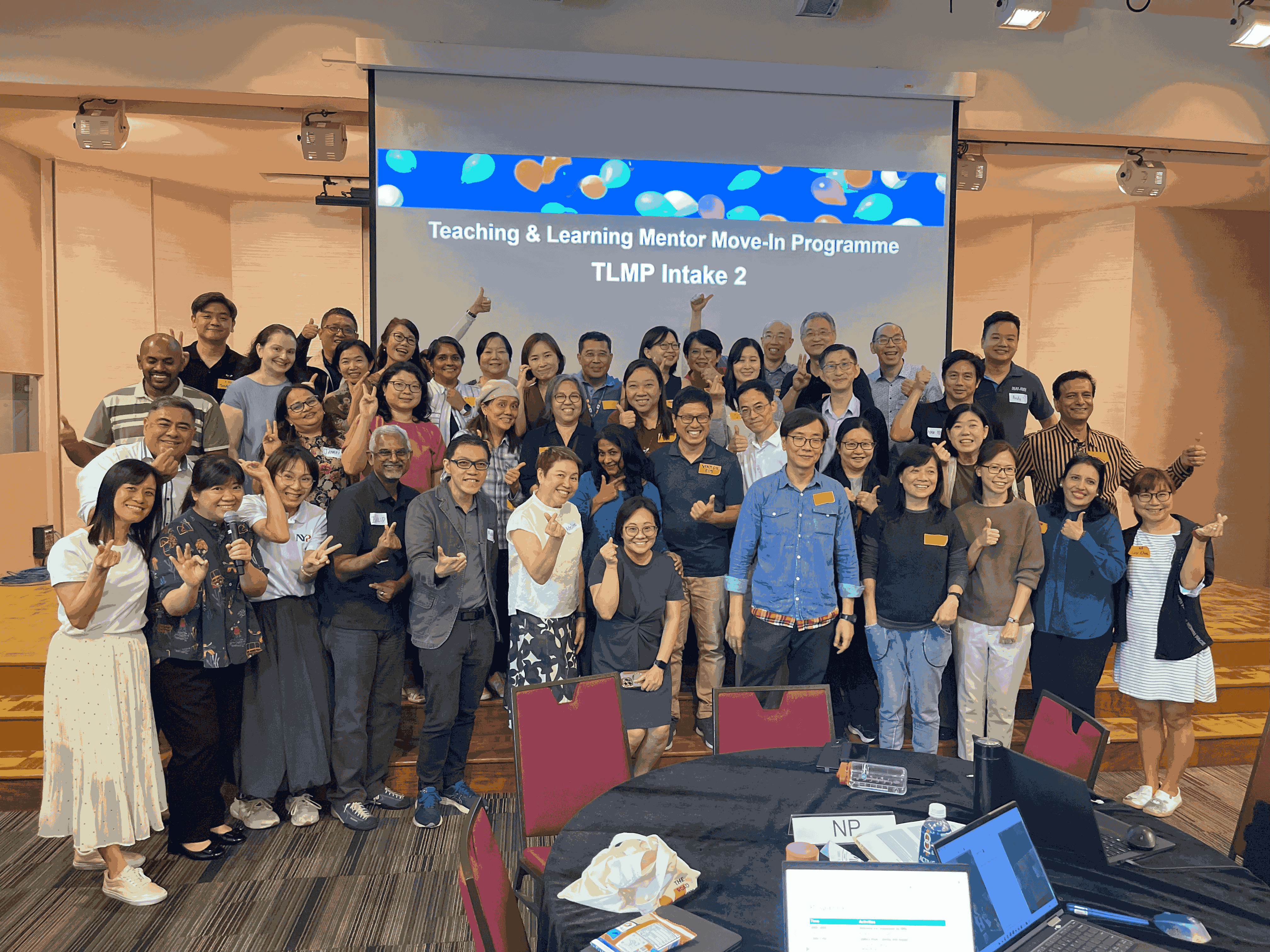 A large group of people posing with thumbs-up gestures in front of a screen displaying "Teaching & Learning Mentor Move-In Programme TLMP Intake 2".