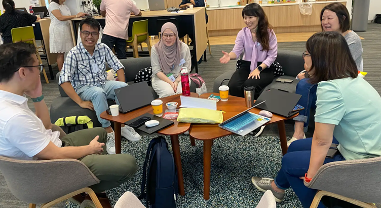 A group of people are seated around a wooden table with laptops, coffee cups, and assorted items in a casual office setting.