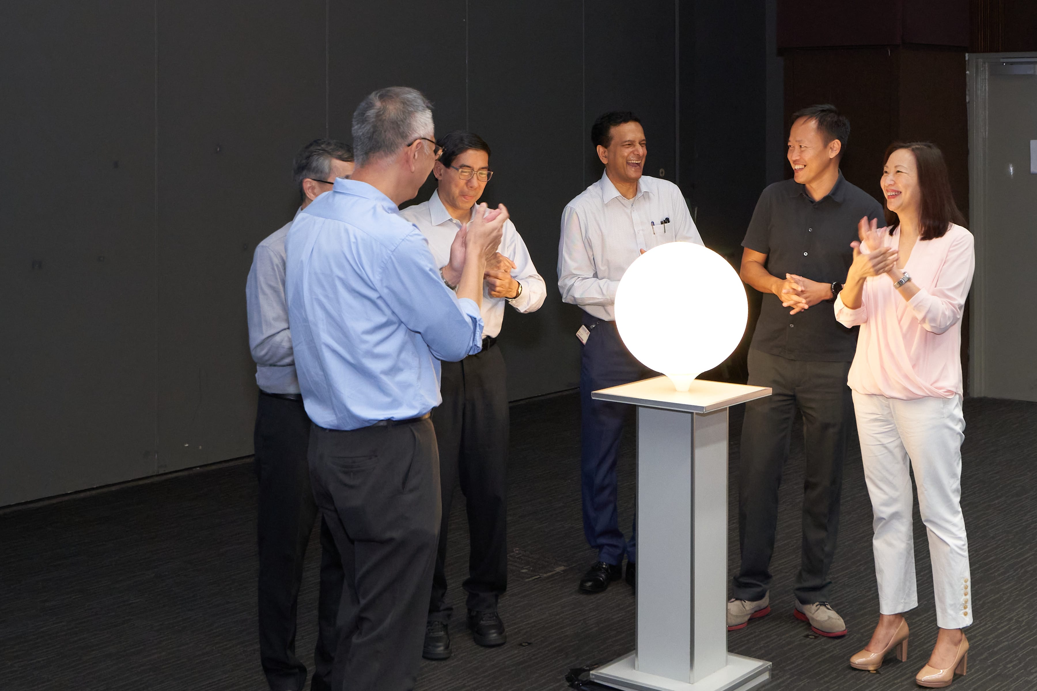 Six people stand in a dark room surrounding a small, pedestal-mounted white orb, which is brightly illuminated. They are all smiling and clapping, appearing to celebrate a launch or lighting moment.