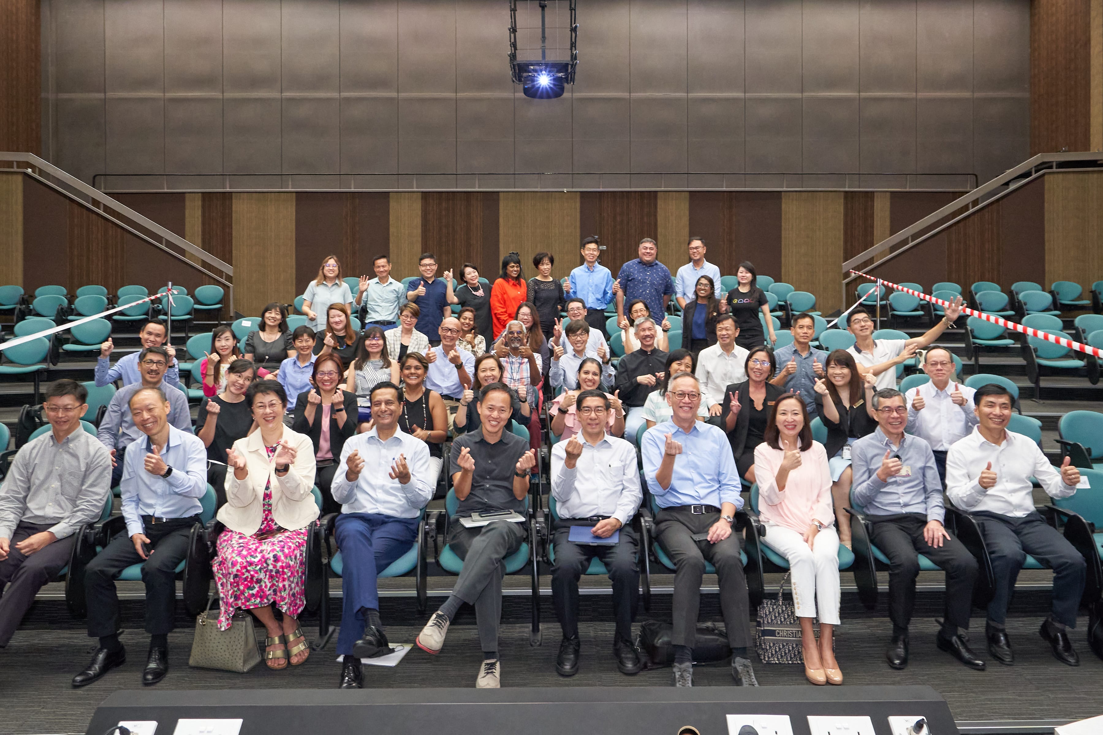 A large group of approximately 50 people are posed together, filling a section of a steep, tiered-seating auditorium. Those in the foreground and a few others are smiling and giving thumbs-up gestures, including one man in the front row who is smiling and crossing his index fingers. The seats behind them are empty.
