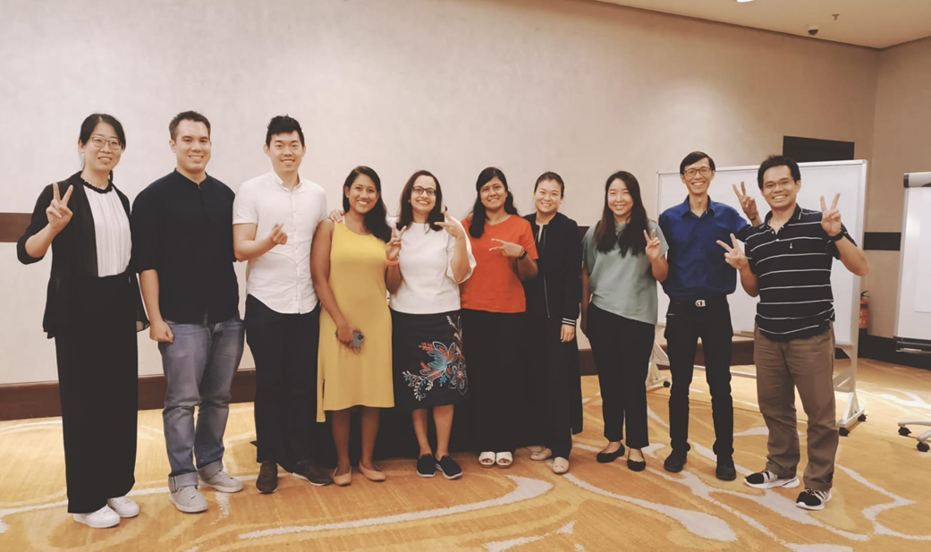 A group of eleven people standing indoors, posing with peace signs and casual attire.