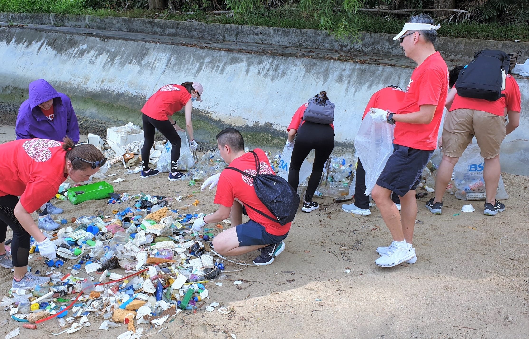 A group of people in red shirts clean up trash on a sandy beach near a concrete wall.