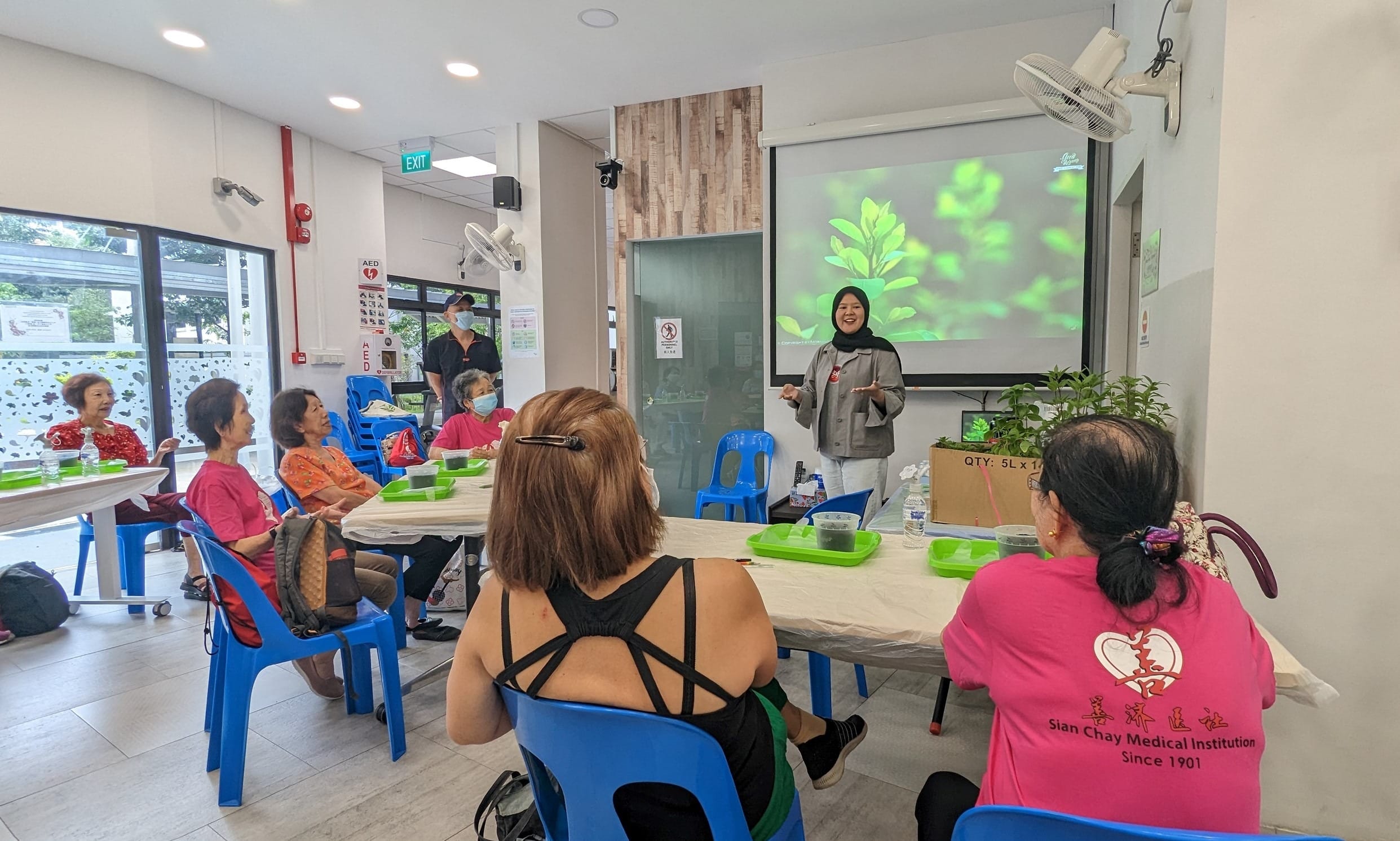 A woman in black headscarf faces a group of people sitting in blue plastic chairs at tables and has a projector screen behind her showing plants. on the tables, there are green trays with items to do a simple transplanting exercise. 
