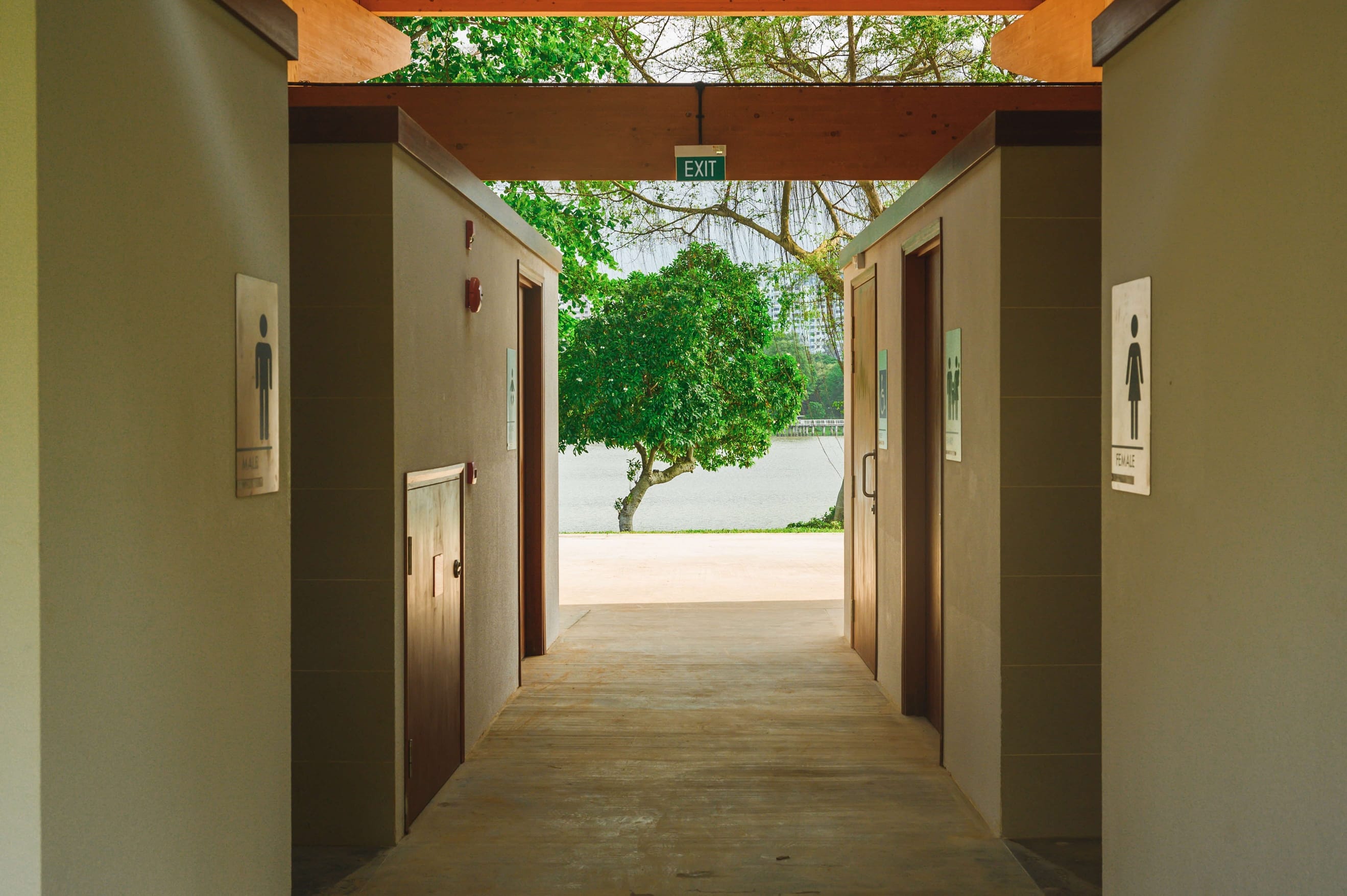 A hallway with male and female restroom signs leads to an outdoor area with green trees and water.