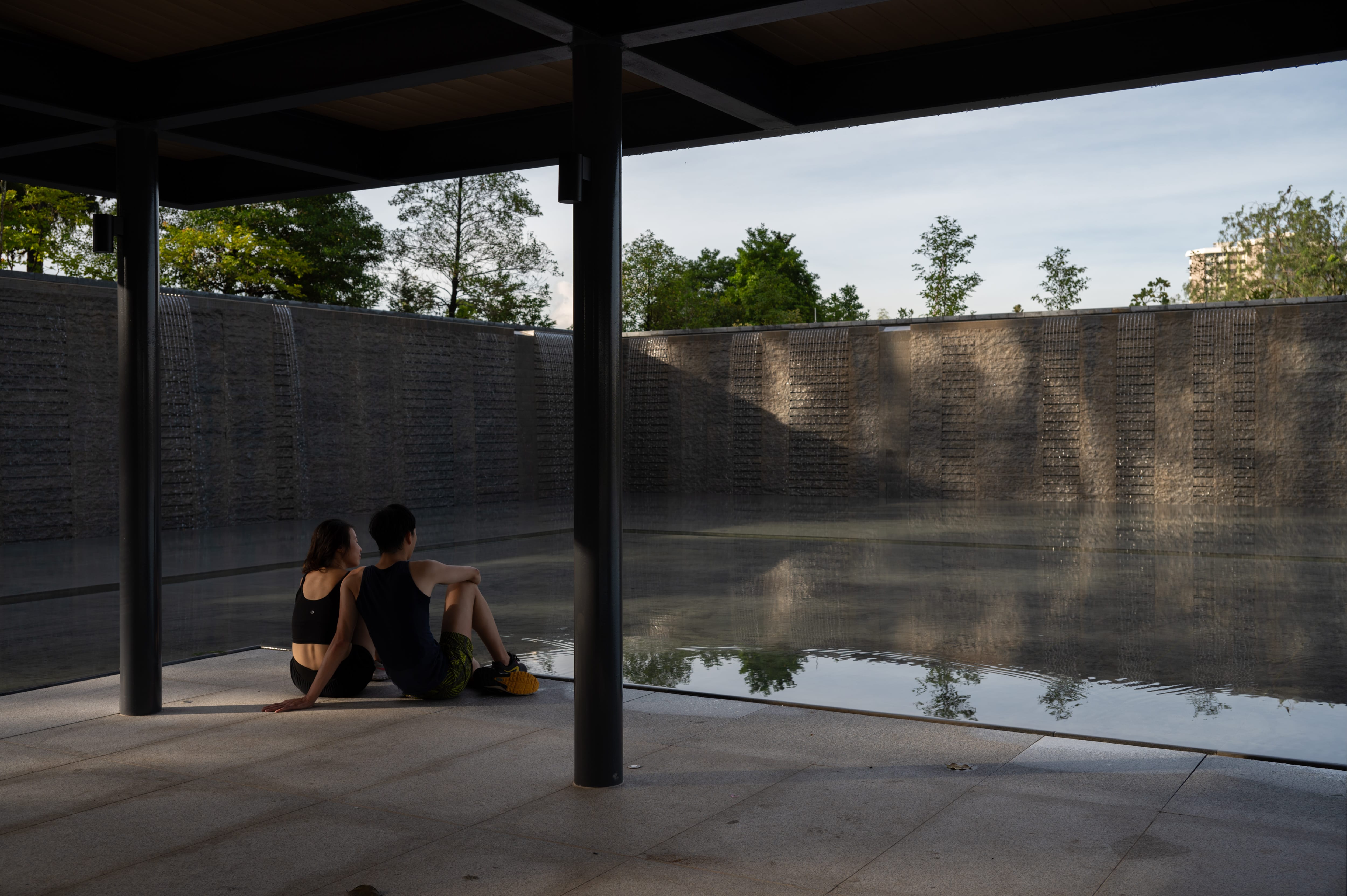 Two individuals sit on the ground near a calm reflective pool with cascading water and trees in the background.
