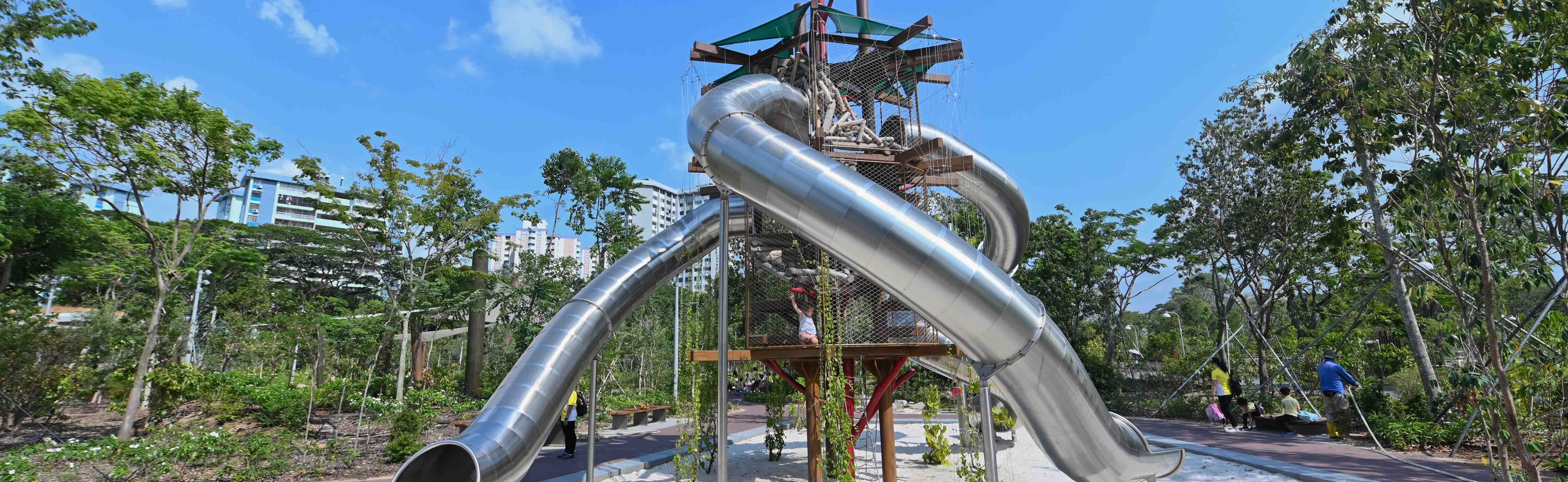 A tall playground structure with large metal slides; trees and buildings are in the background.