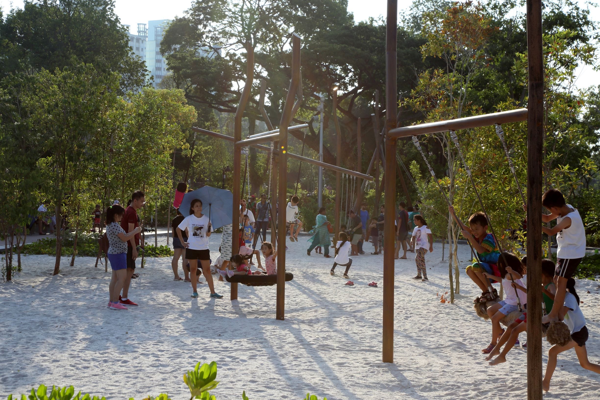 Children and adults enjoy a sunny day in a playground with swings in a sandy area surrounded by trees.