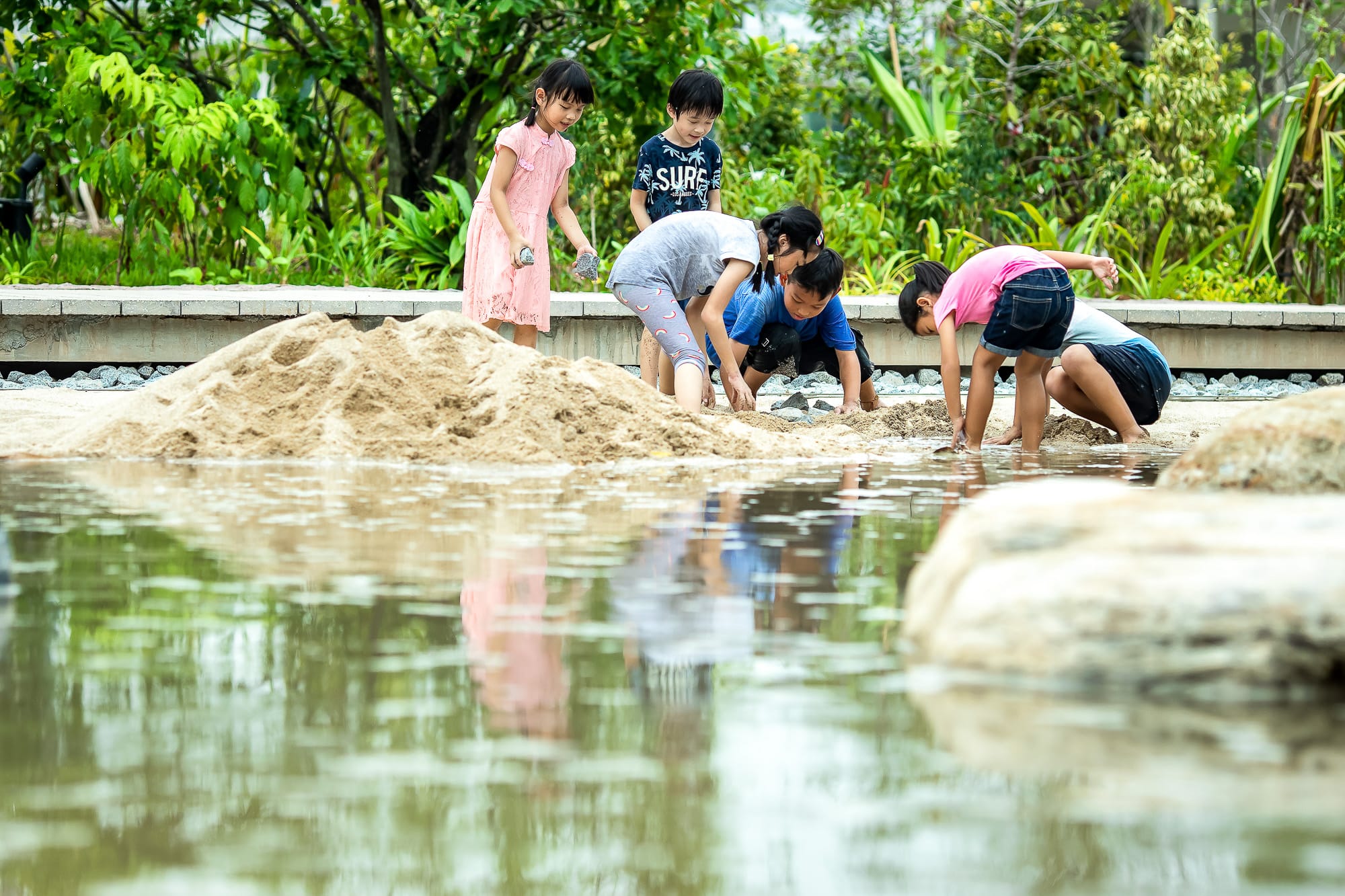 Several children are playing with sand beside a shallow water body in a lush garden with green plants in the background.