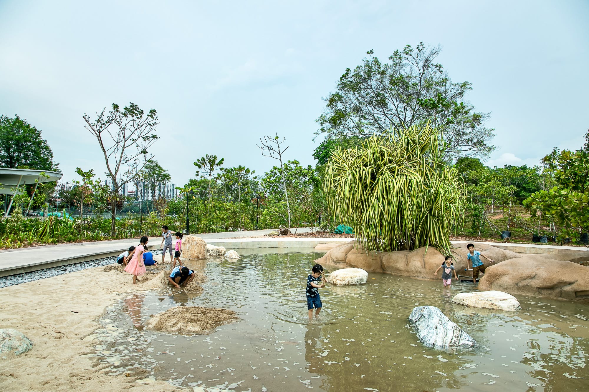 Children playing in sandy and shallow water near trees and large rocks in an outdoor park on a cloudy day.