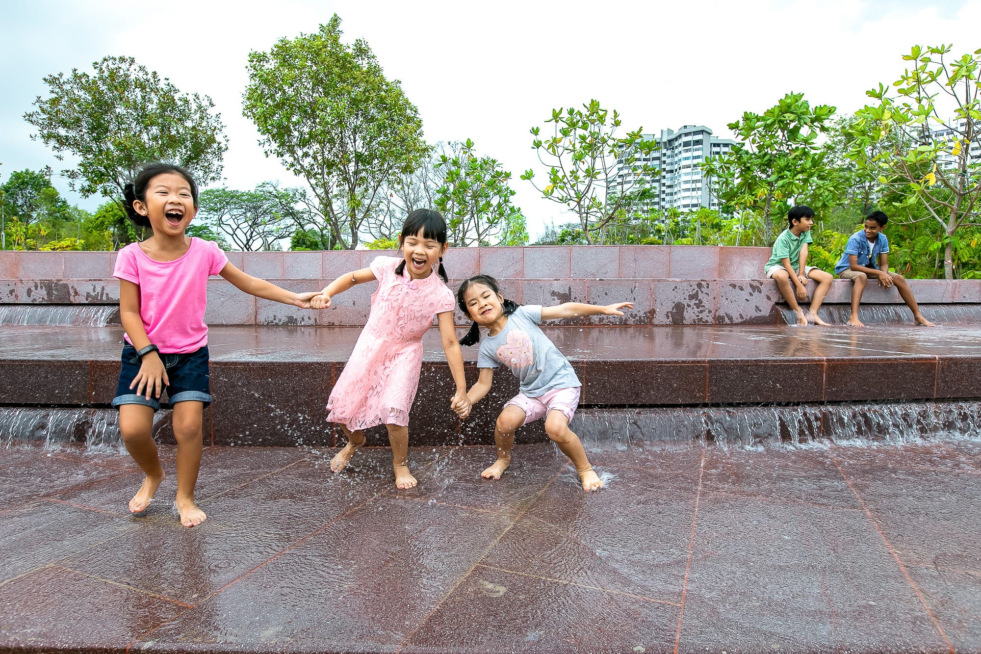 Three children play in a water feature while two others sit on steps; trees and buildings are visible in the background.
