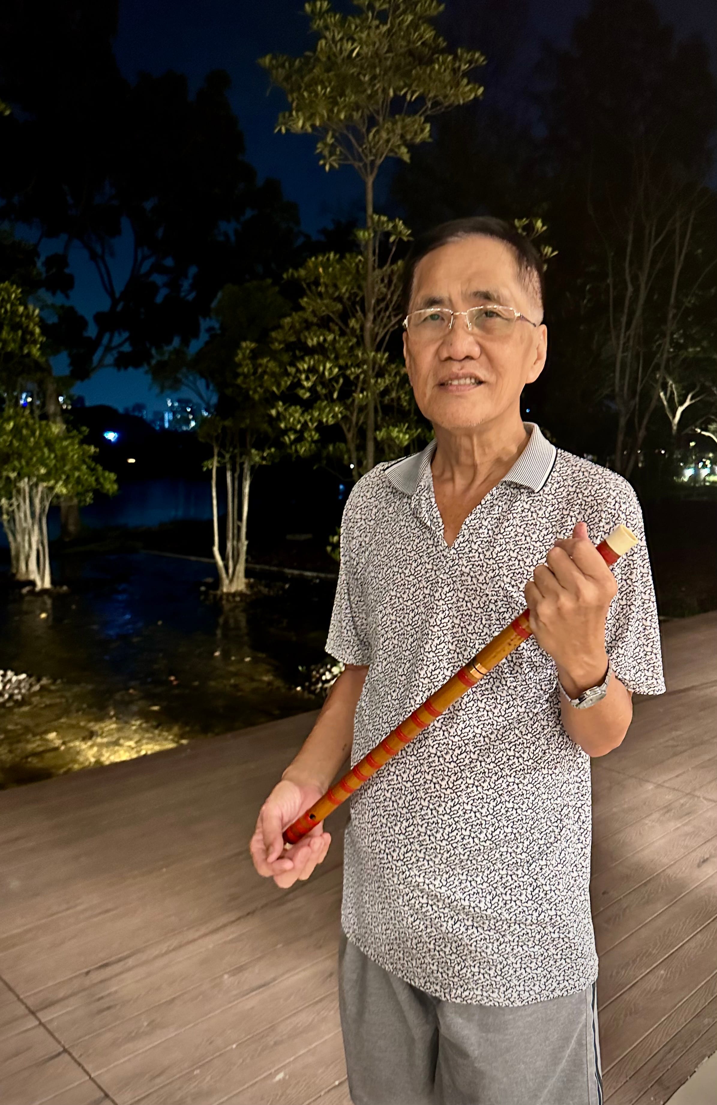 A person wearing a grey patterned shirt holds a wooden stick outdoors at night, surrounded by trees and water.