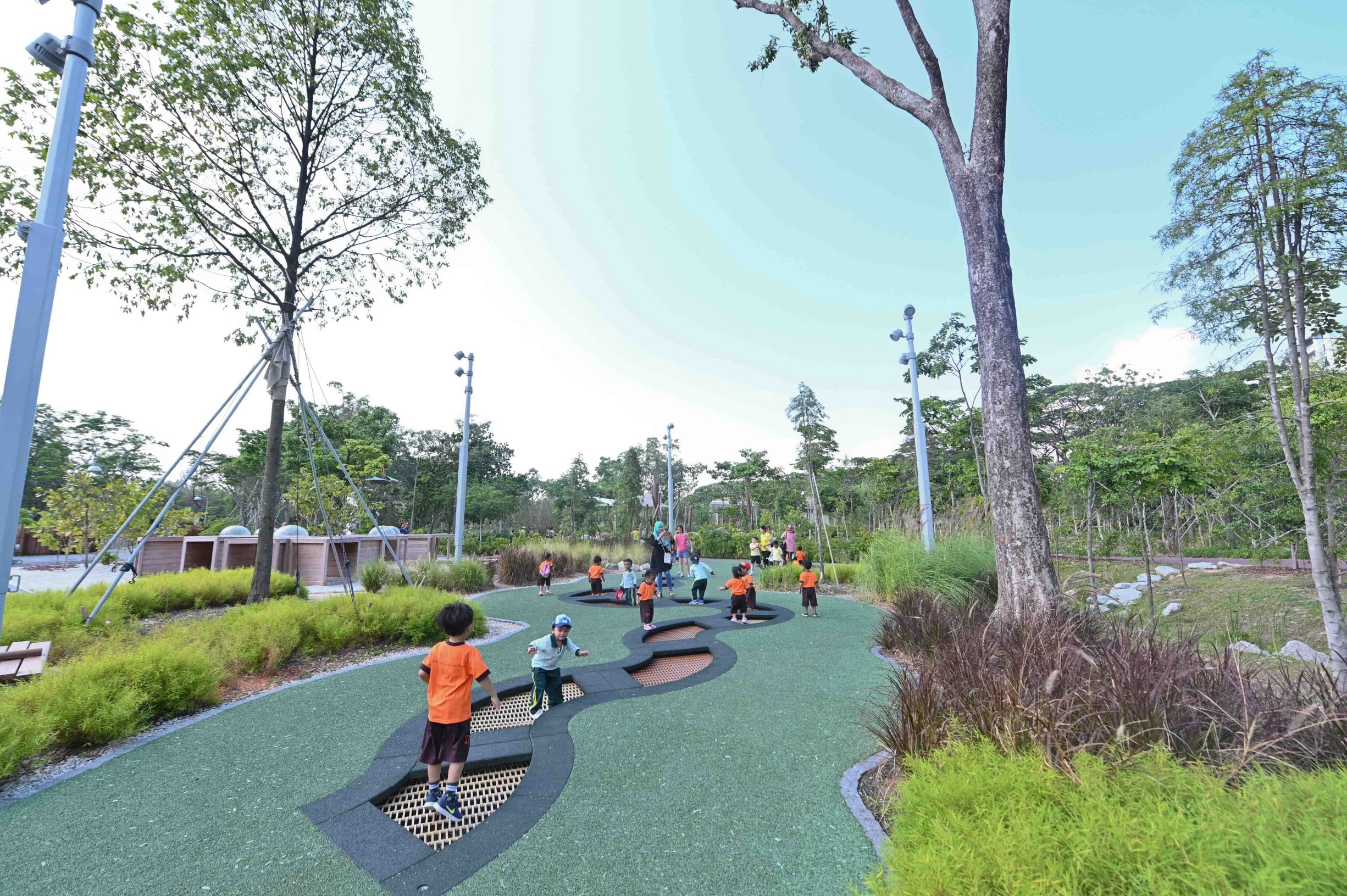 Children play on green pathways near trees and playground equipment in a park under a clear blue sky.