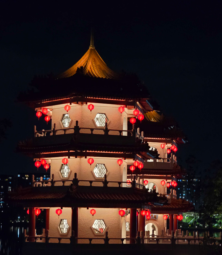 A multi-tiered Chinese pagoda illuminated with red lanterns at night, with a cityscape faintly visible in the background.