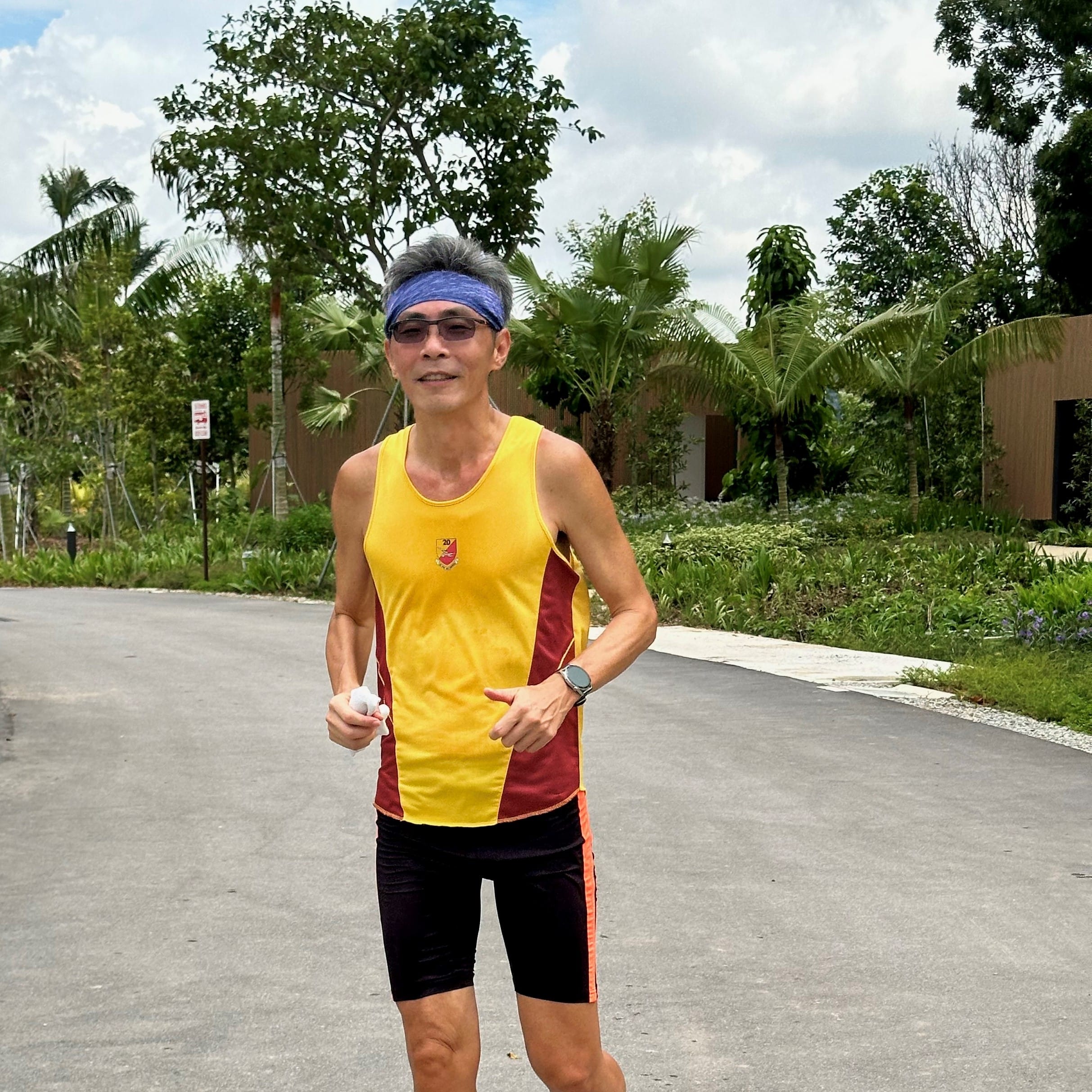 A person wearing a yellow and red athletic tank top and black shorts is jogging outside near greenery on a paved path.
