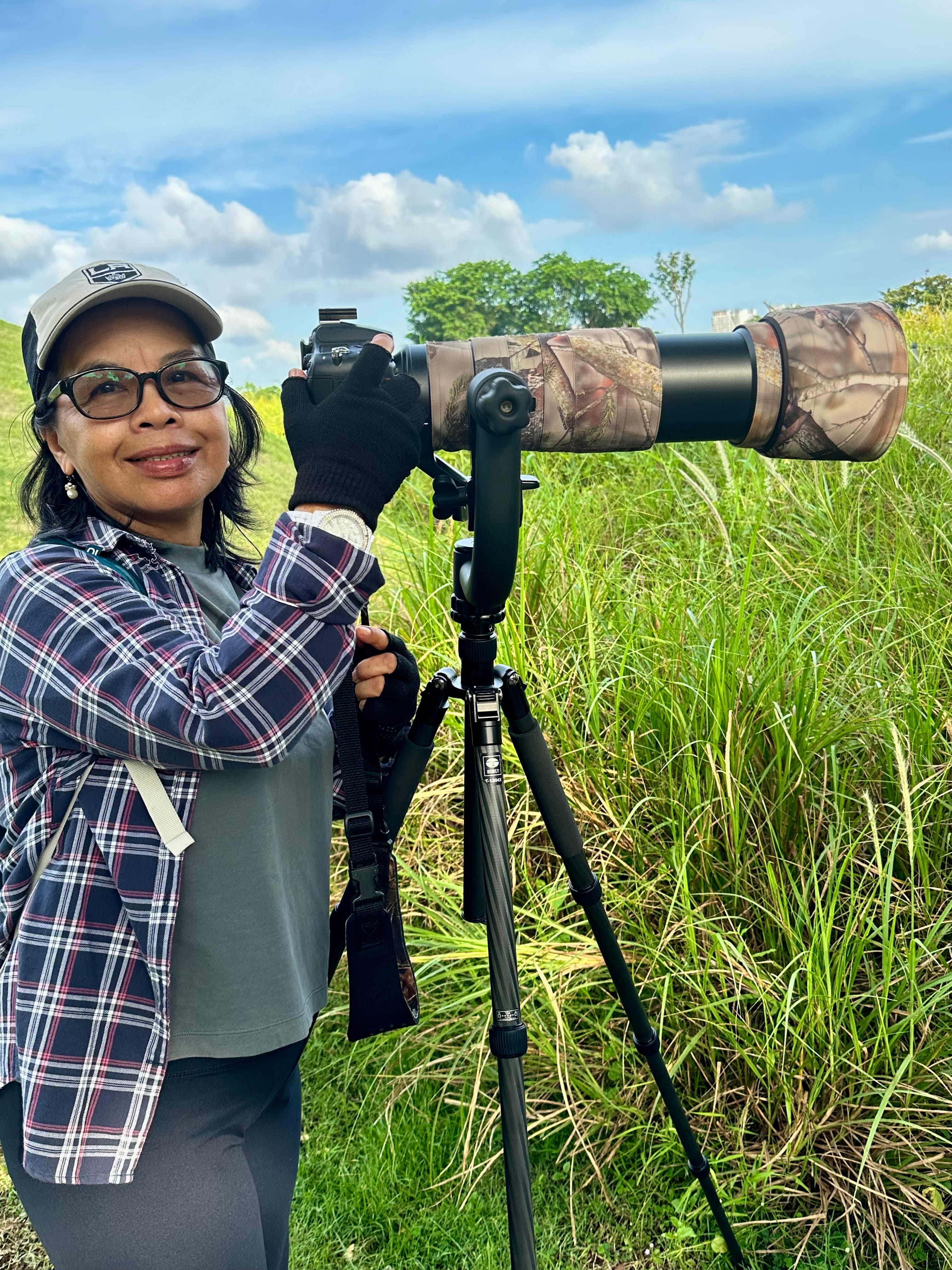 A person wearing a plaid shirt uses a camera with a large camouflaged lens on a tripod in a grassy field.