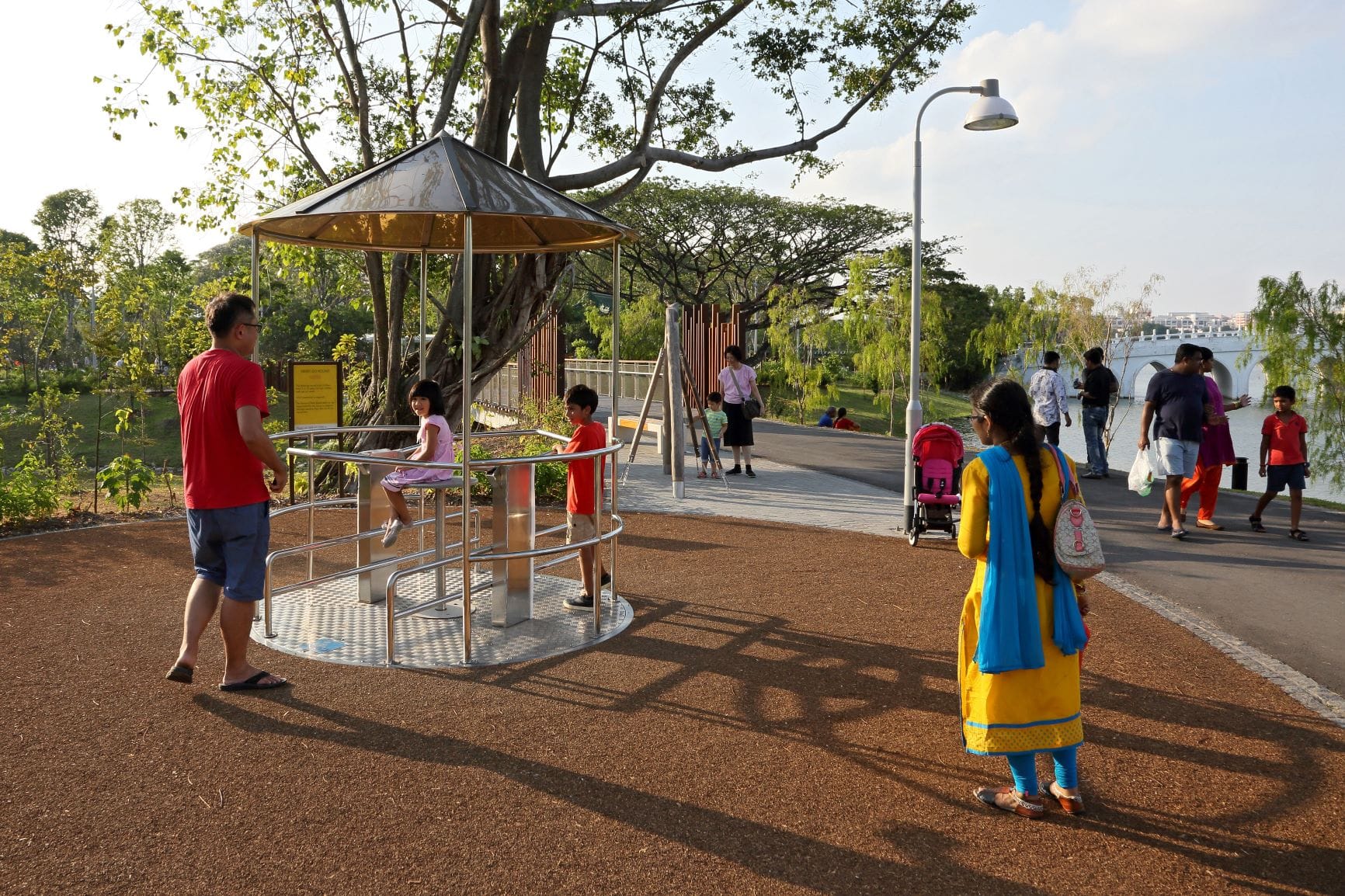A playground with children playing, surrounded by adults, trees, and a paved pathway next to a body of water.