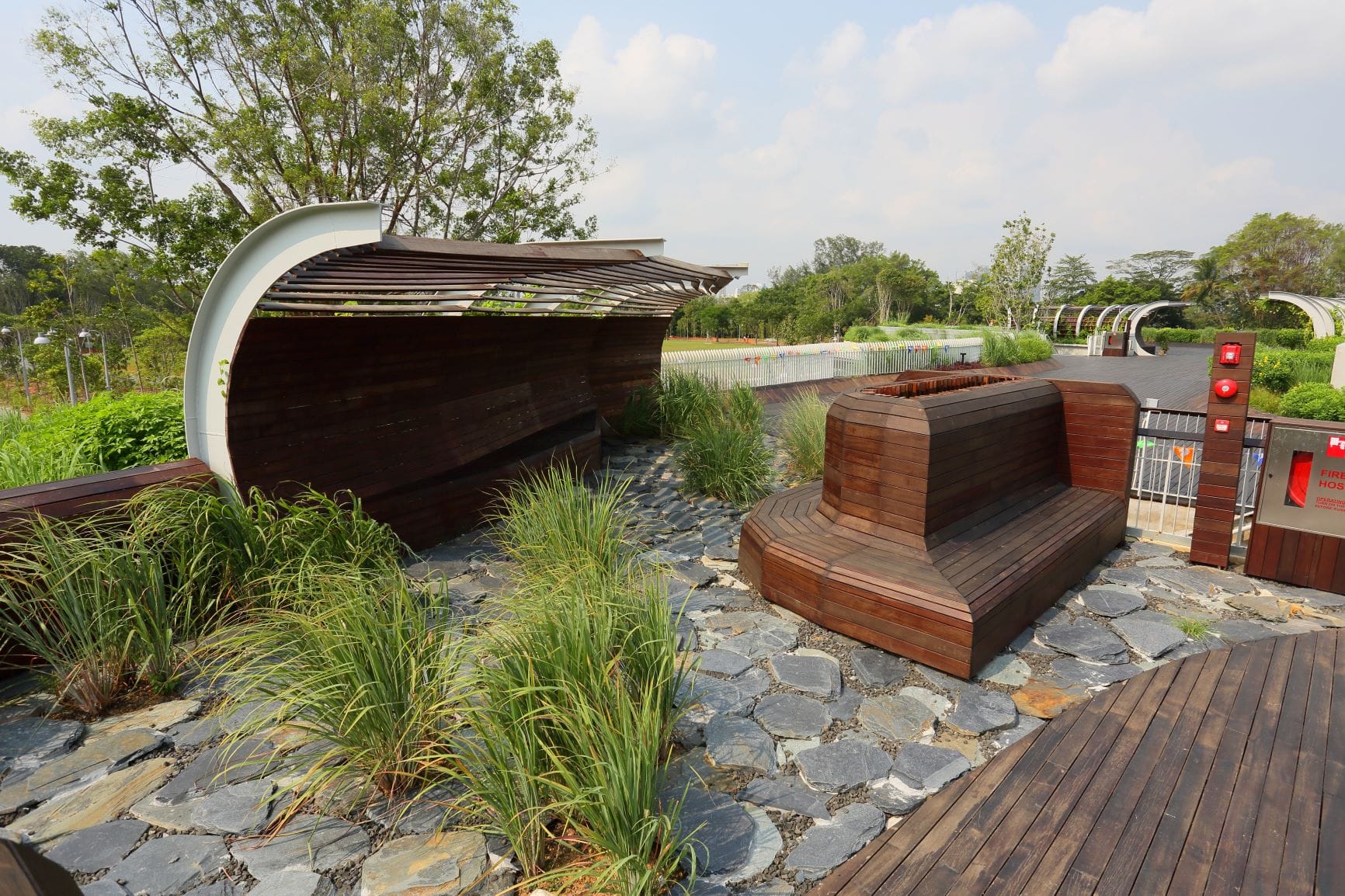 A curved wooden bench with a canopy, surrounded by plants and stone tiles, in a park setting under a partly cloudy sky.