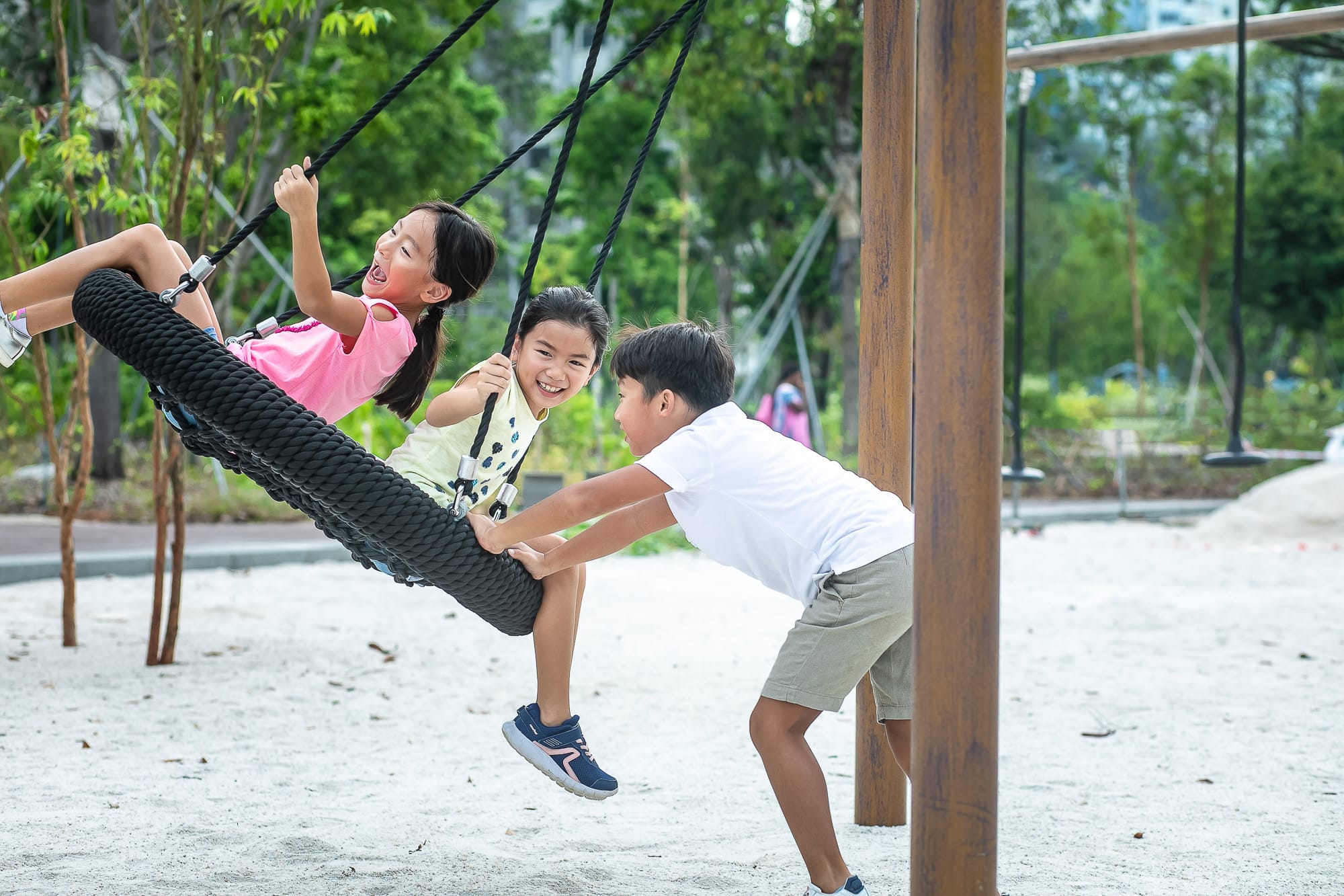 Three children are playing on a tire swing in a sandy playground, surrounded by trees and greenery.