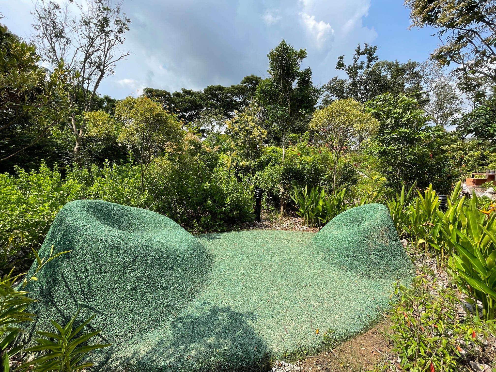 A green grassy structure surrounded by dense, green trees and plants under a partly cloudy sky.