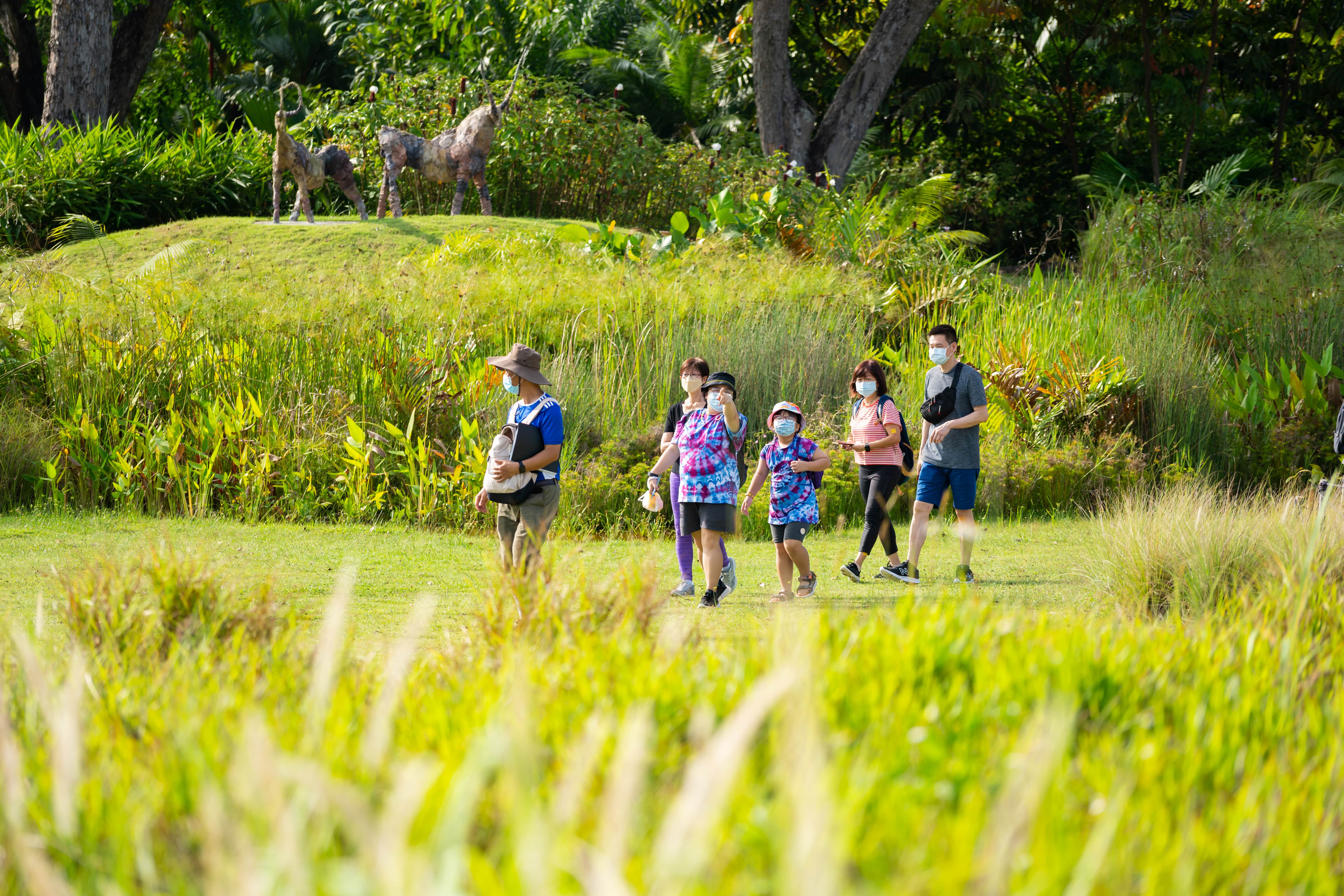 Several people walk through a grassy area with two giraffe sculptures visible in the background.