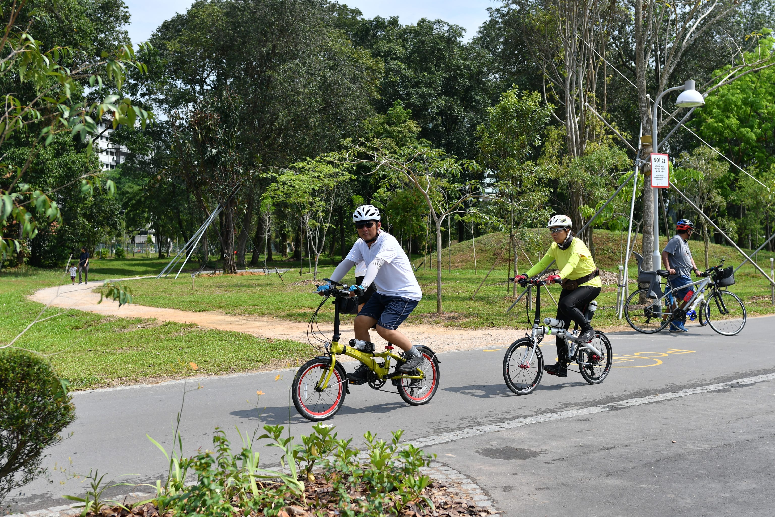 Two cyclists ride along a paved path in a park surrounded by trees, while a pedestrian walks in the background.