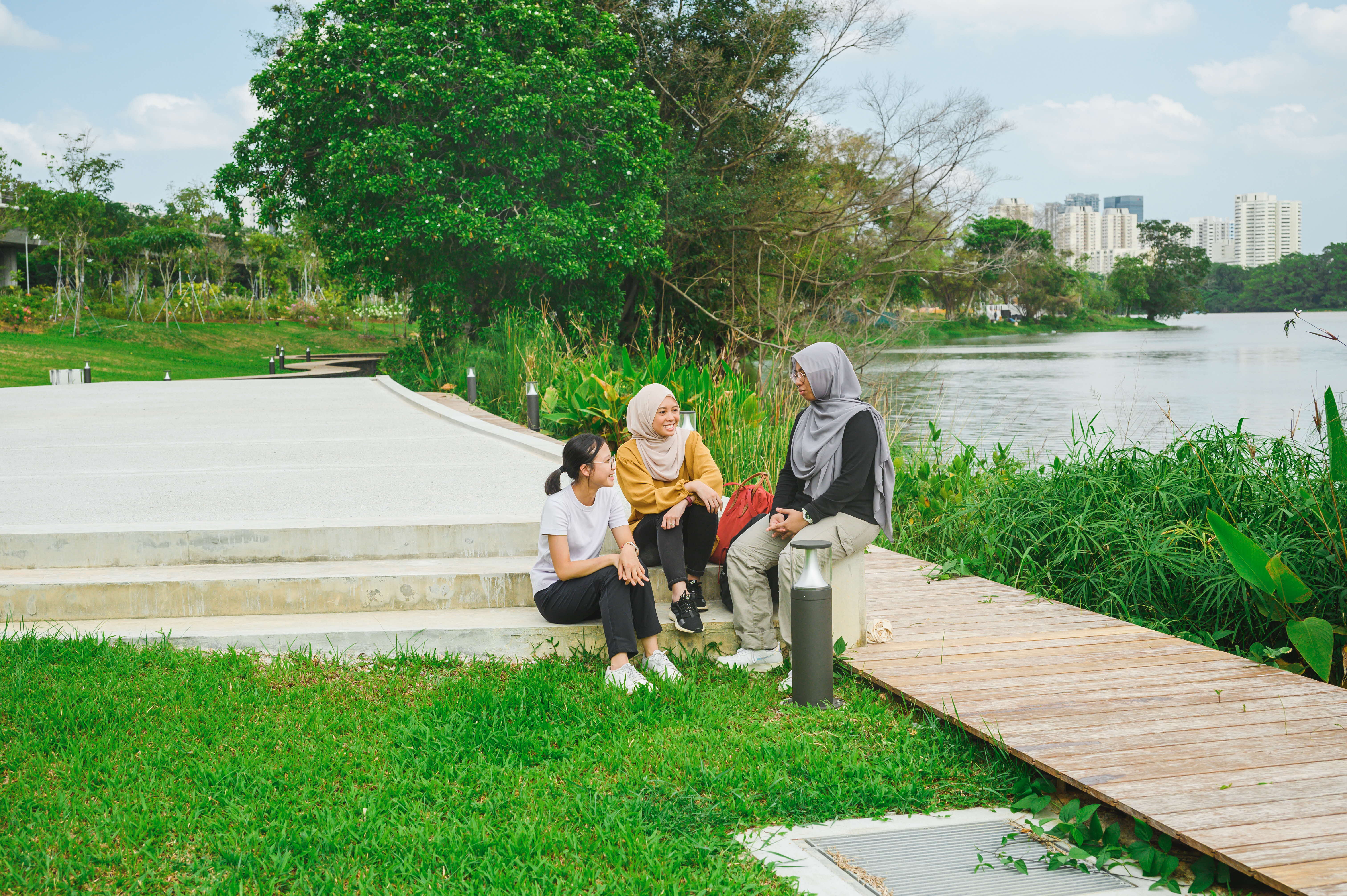Three people sit and converse in a green park next to a river with buildings visible in the background.