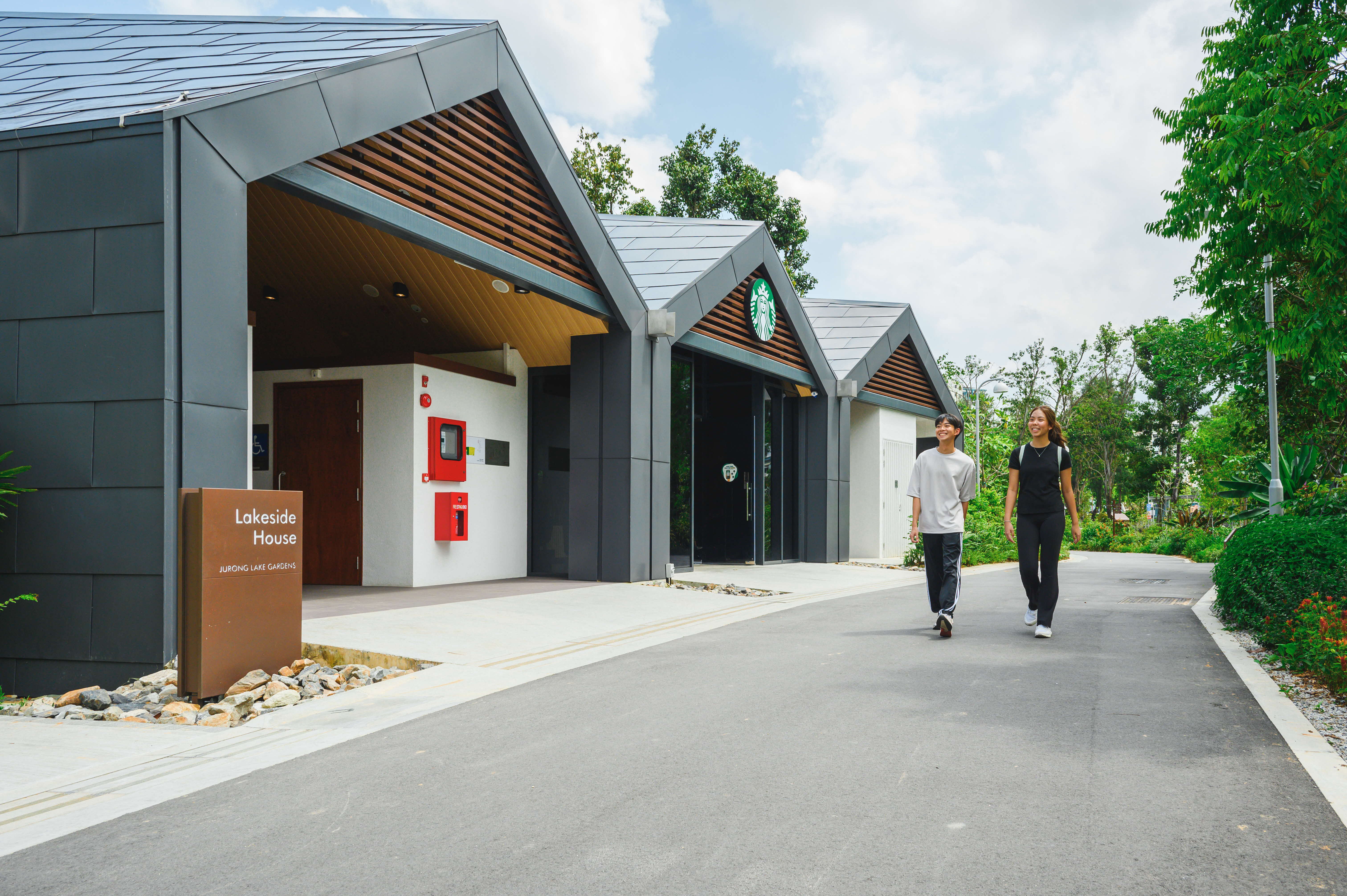 Two people walking on a pathway beside Lakeside House and a Starbucks with greenery and trees in the background.