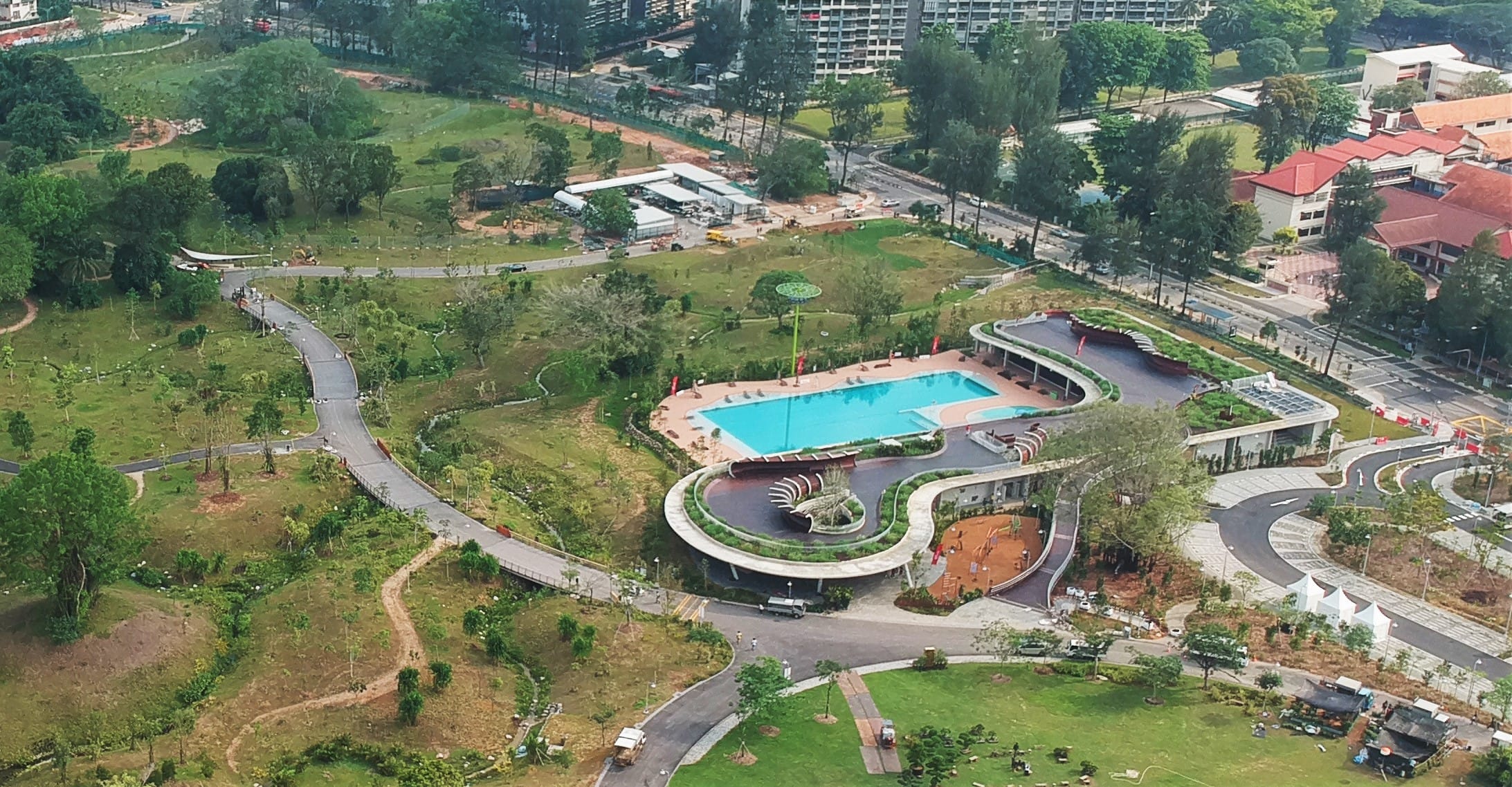 Aerial view of a park with winding paths, trees, a swimming pool, buildings, and surrounding roads.