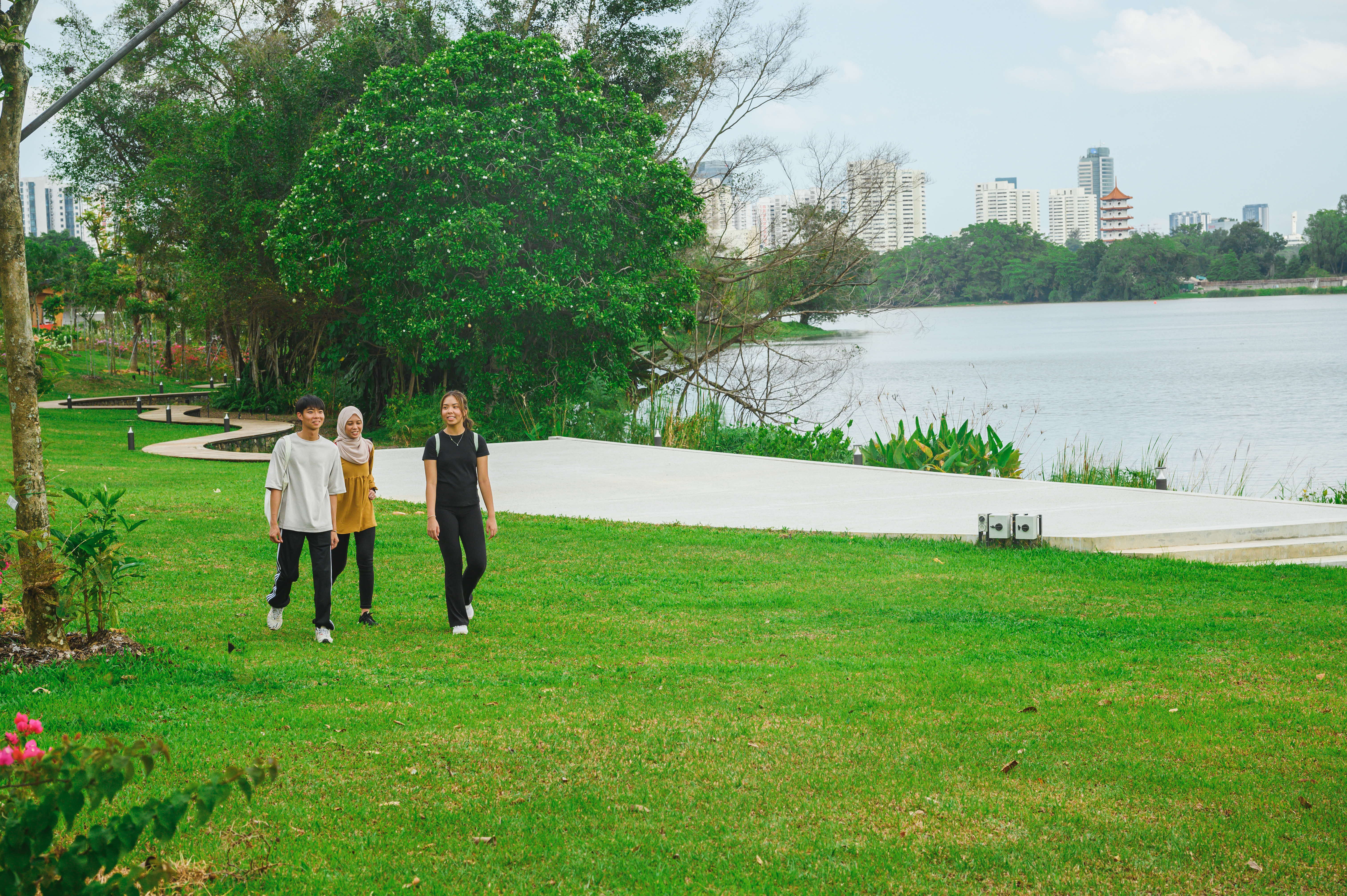 Three people walk on a grassy field beside a lake with lush trees and city buildings in the background.