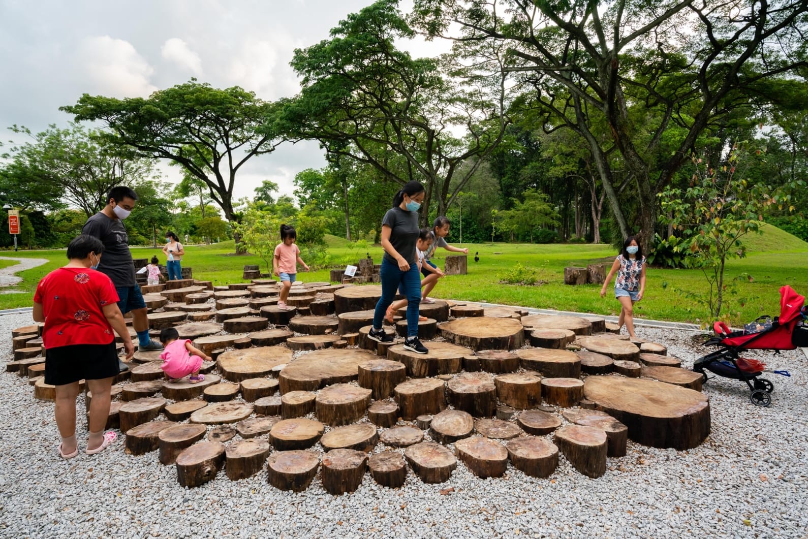 People, including children, are playing on large tree stump slices in a park with lush greenery and gravel paths.