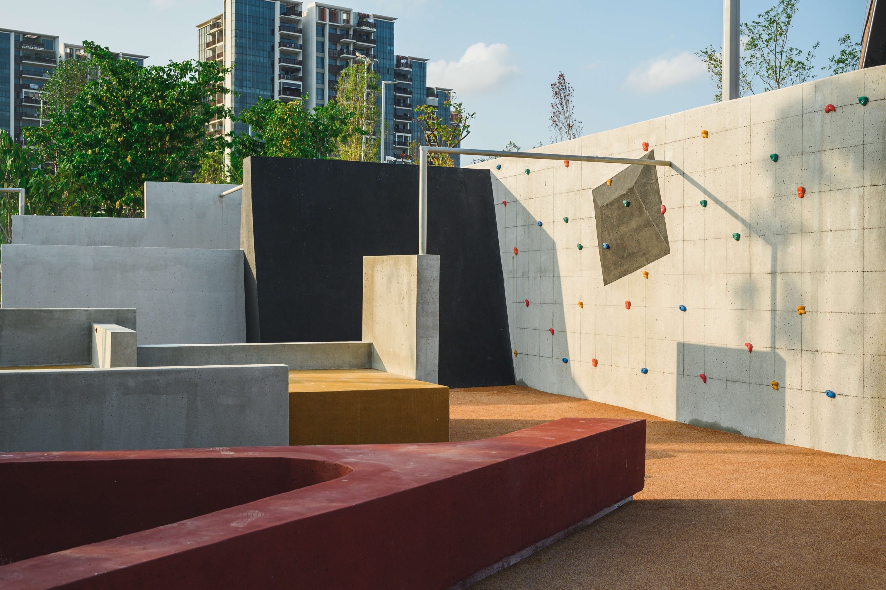 Concrete structures and a climbing wall with colorful grips in an outdoor area, with buildings and trees in the background.