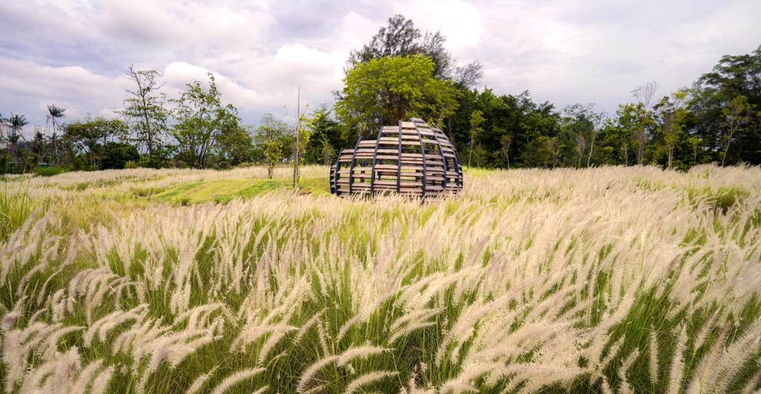 A round wooden structure stands in a grassy field, surrounded by tall, white feathery plants and trees under a cloudy sky.