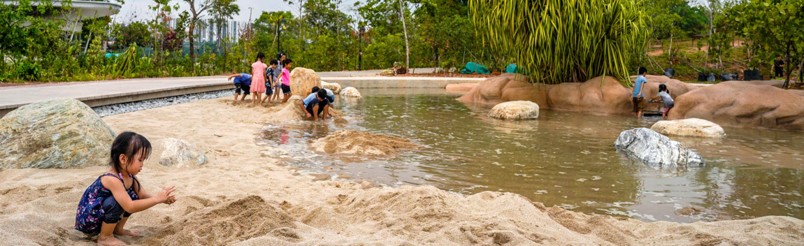 Children play in sand and shallow water at a natural park with trees in the background.