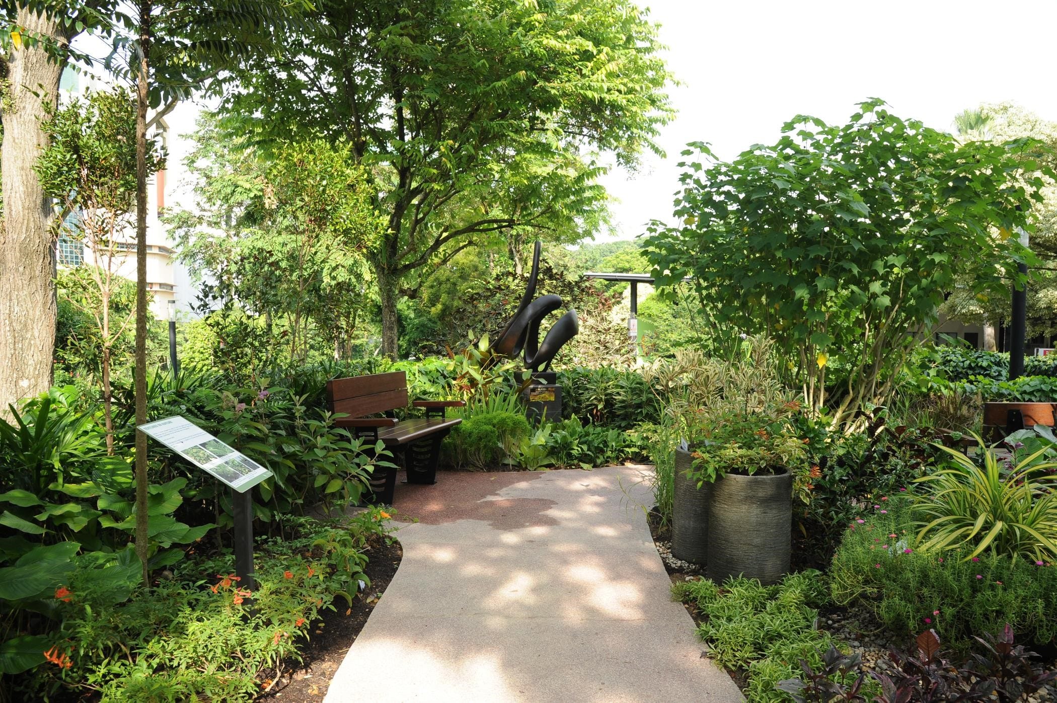 A park with lush greenery, a pathway, benches, a sculpture, and informational sign amidst various plants and trees.