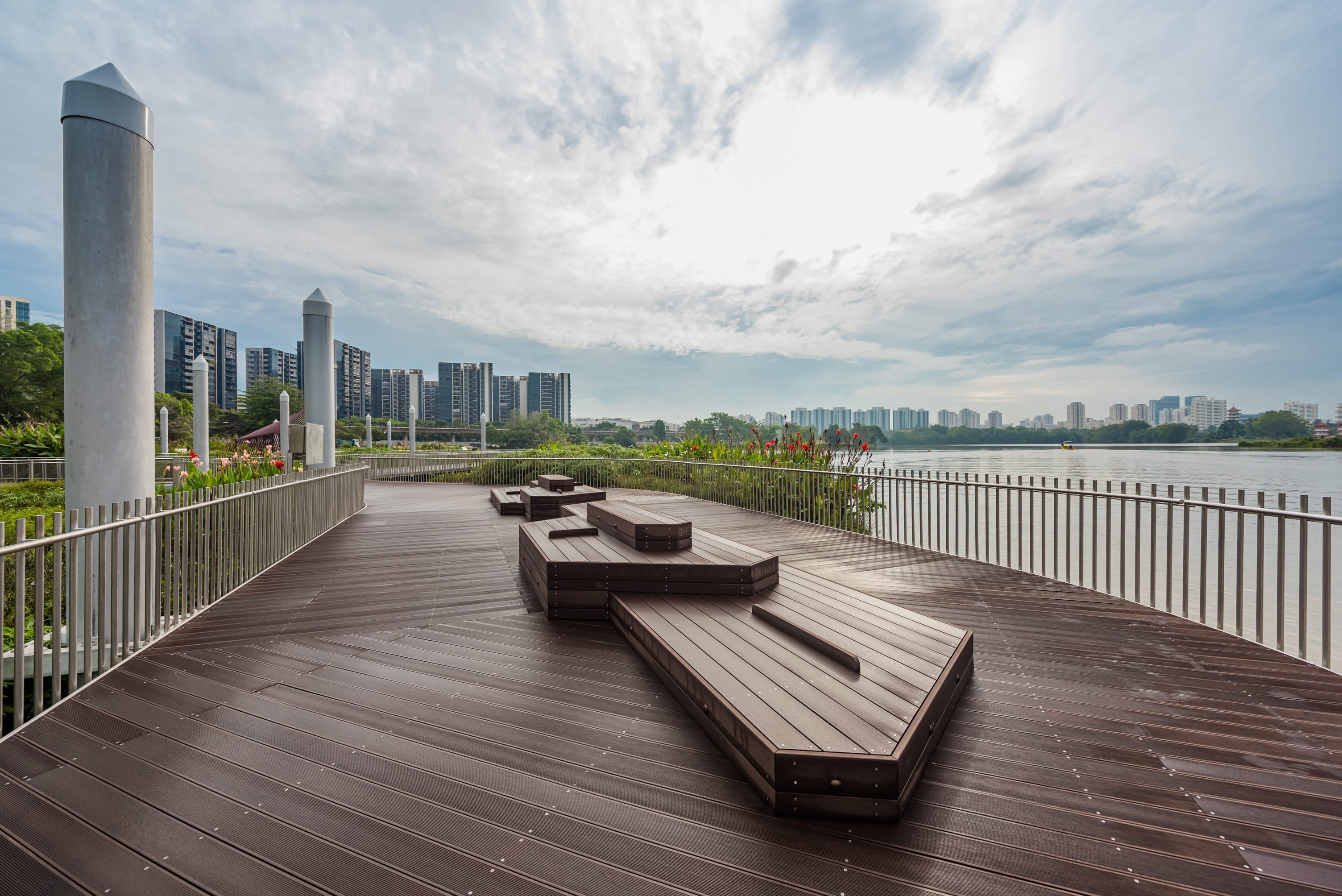 Wooden deck by a body of water with benches, garden, columns, and modern buildings in the background.