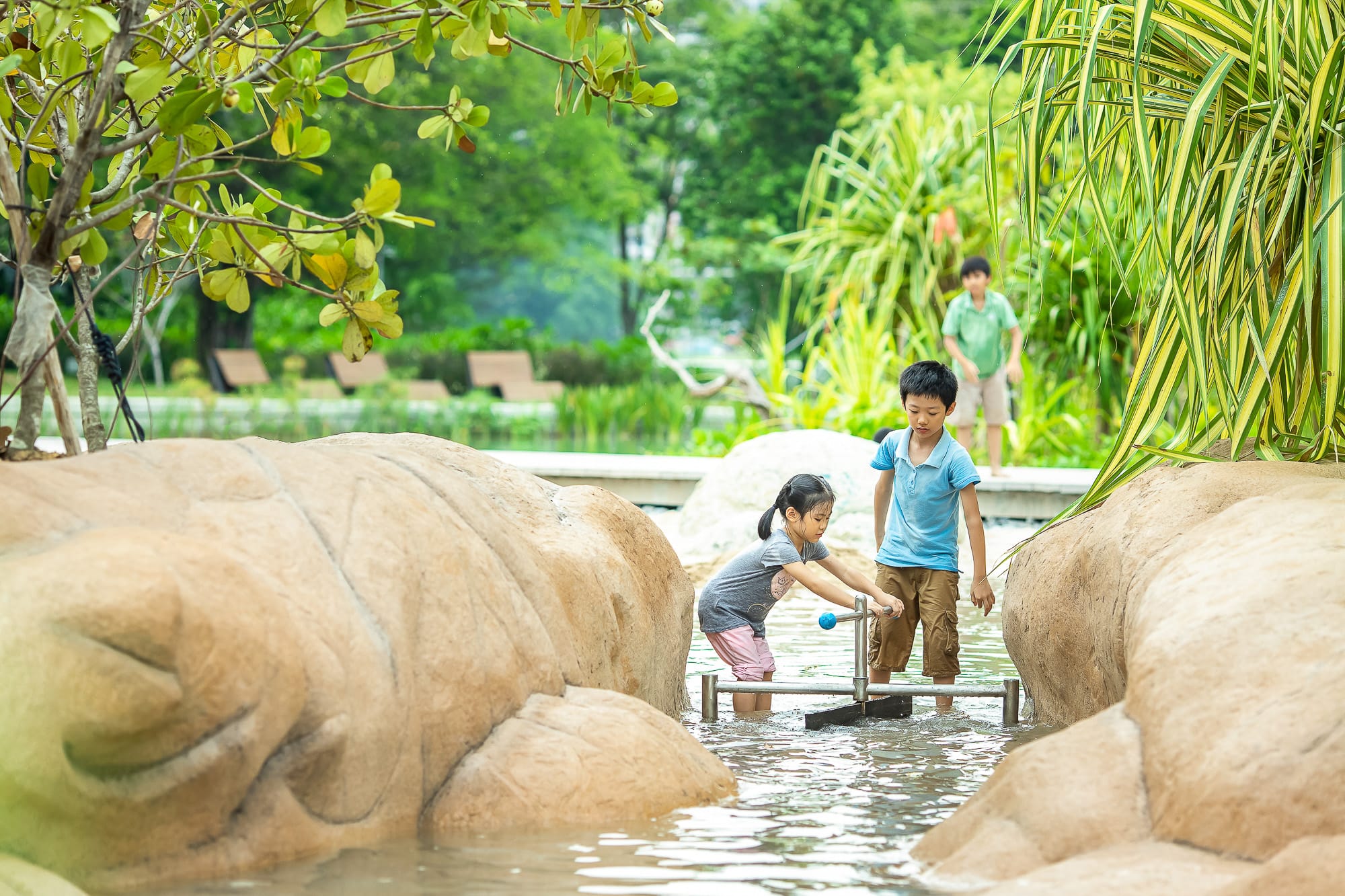 Two children play with a water pump in a shallow stream surrounded by rocks and greenery.