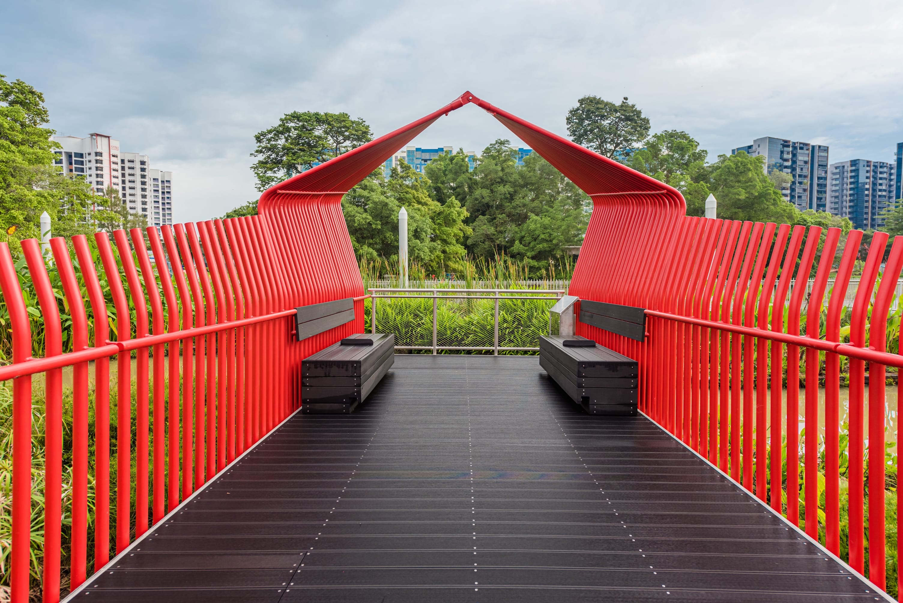 A red pavilion with black benches on a walkway overlooks greenery with buildings in the background.