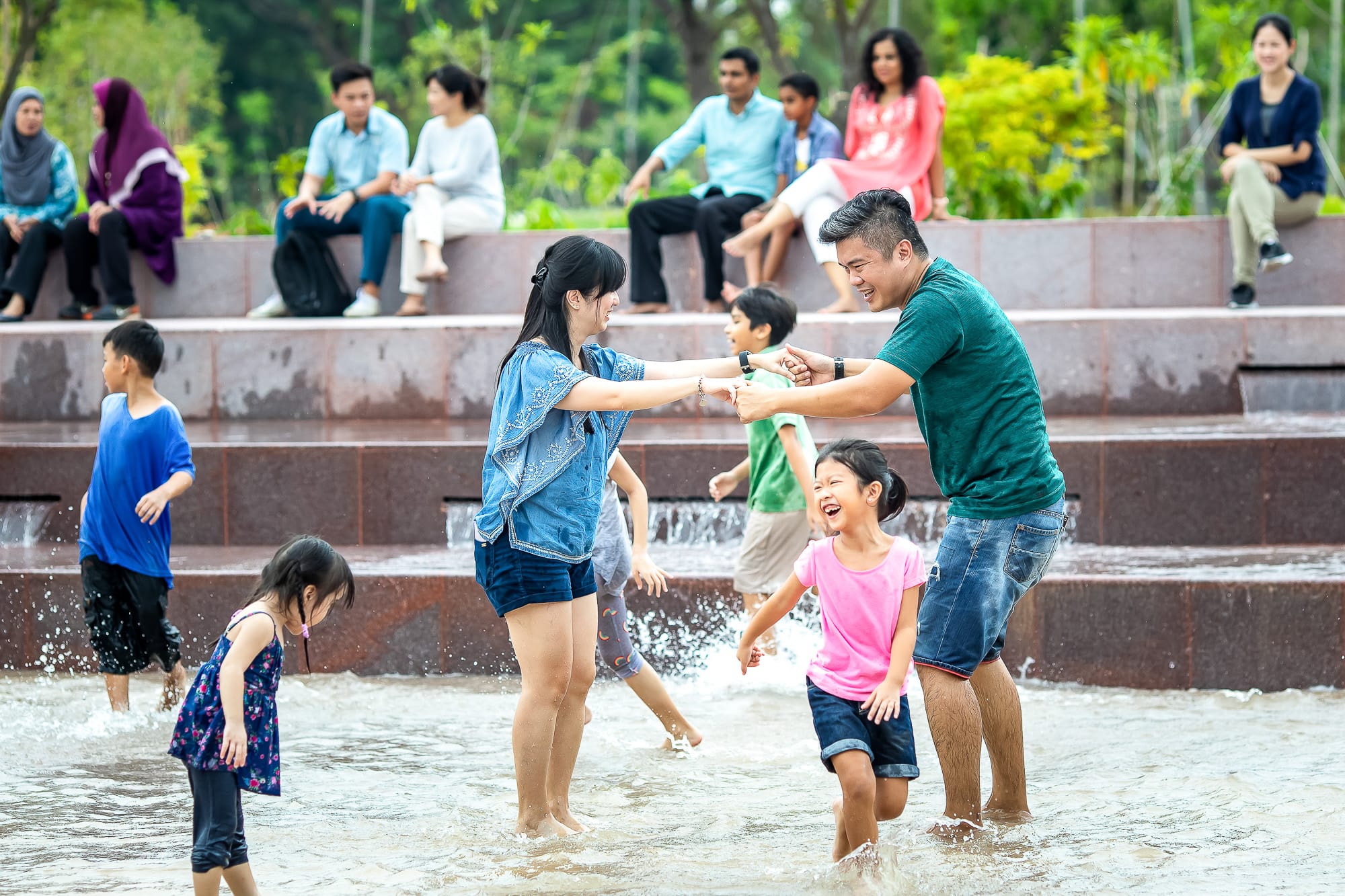 Adults and children play in knee-deep water near tiered stone steps; others sit on the steps and observe.