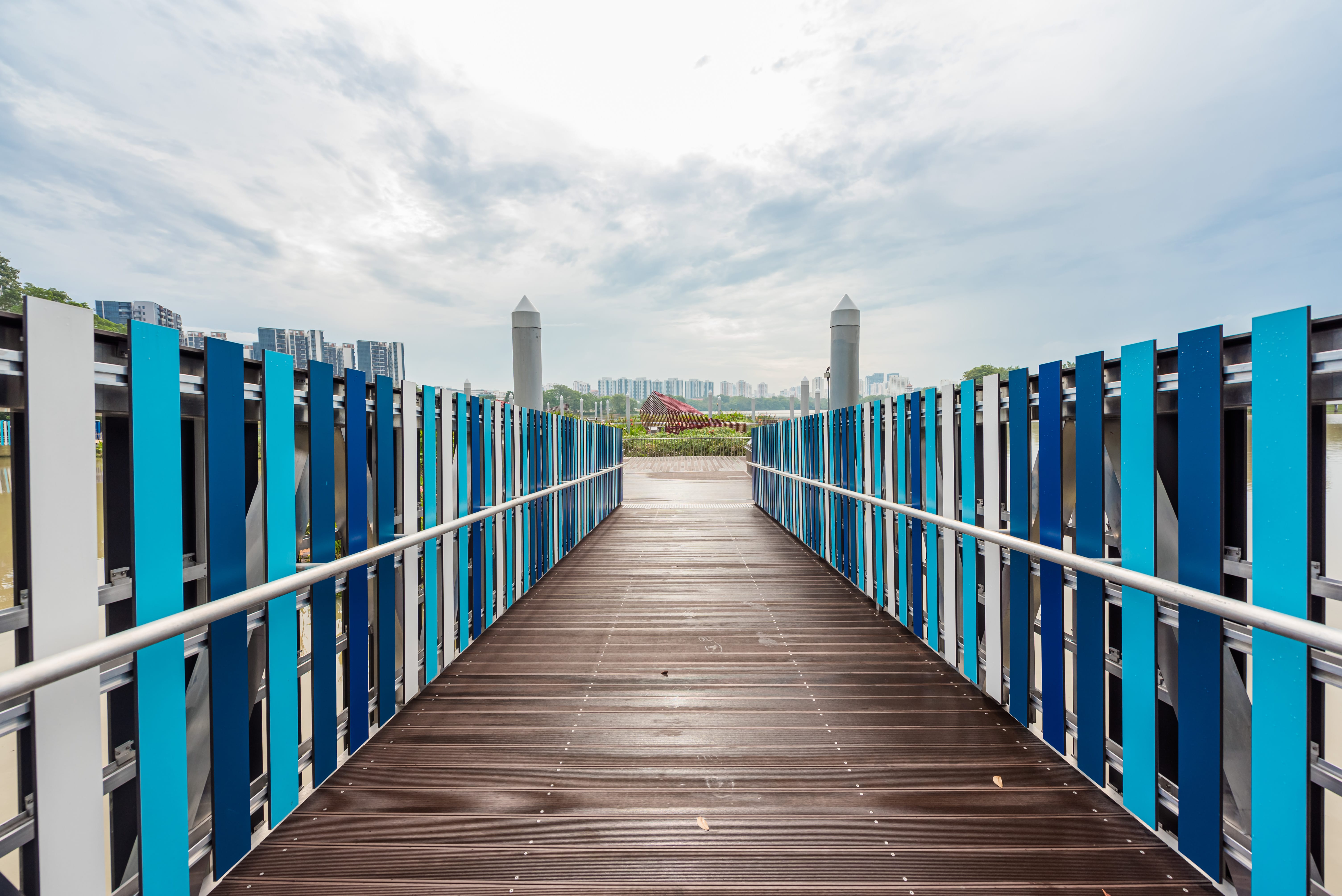 A wooden bridge with blue and white railings leading to a park, with city buildings visible in the background.