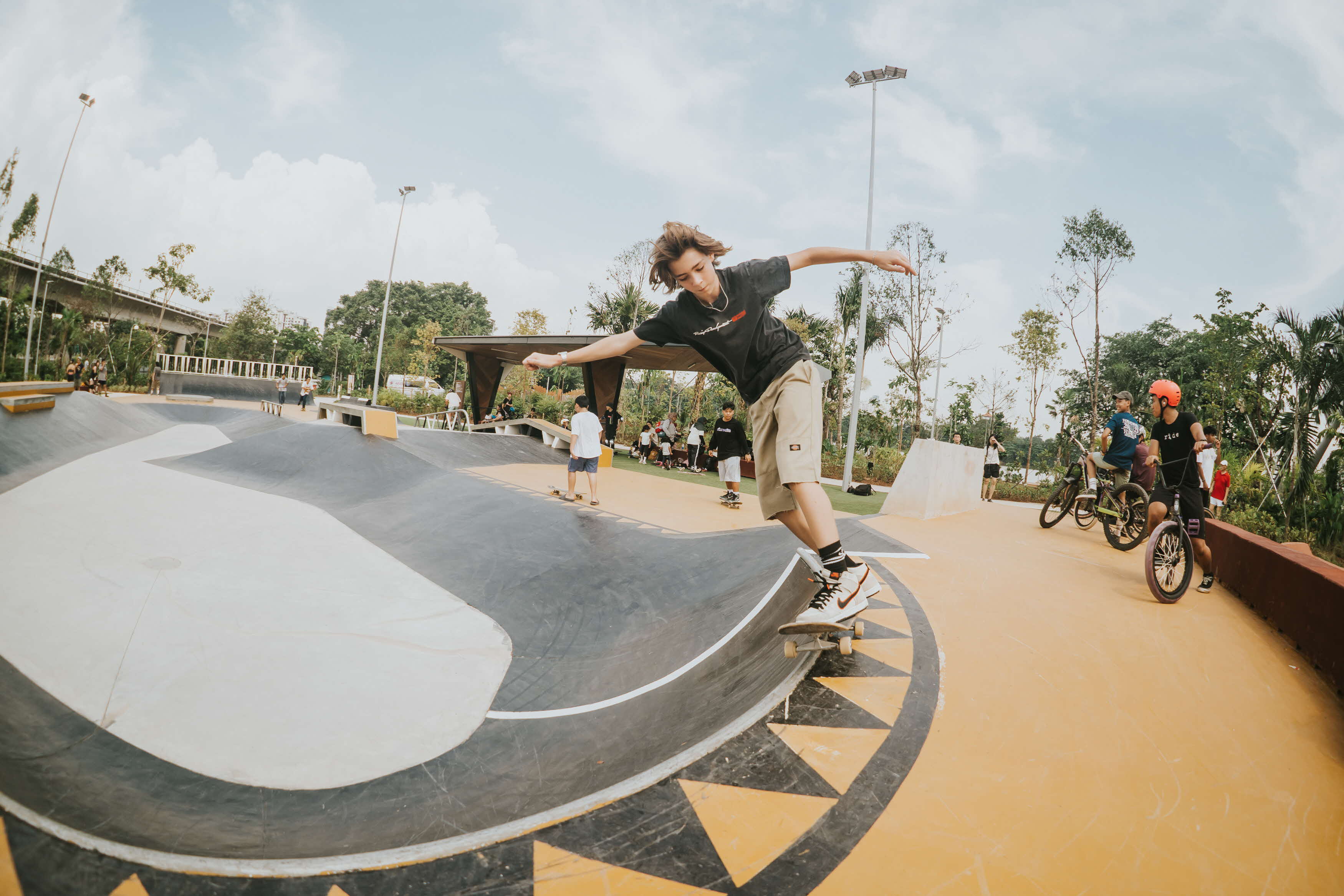 A skateboarder is doing a trick on a ramp in a skatepark, with several other people watching and riding bikes nearby.