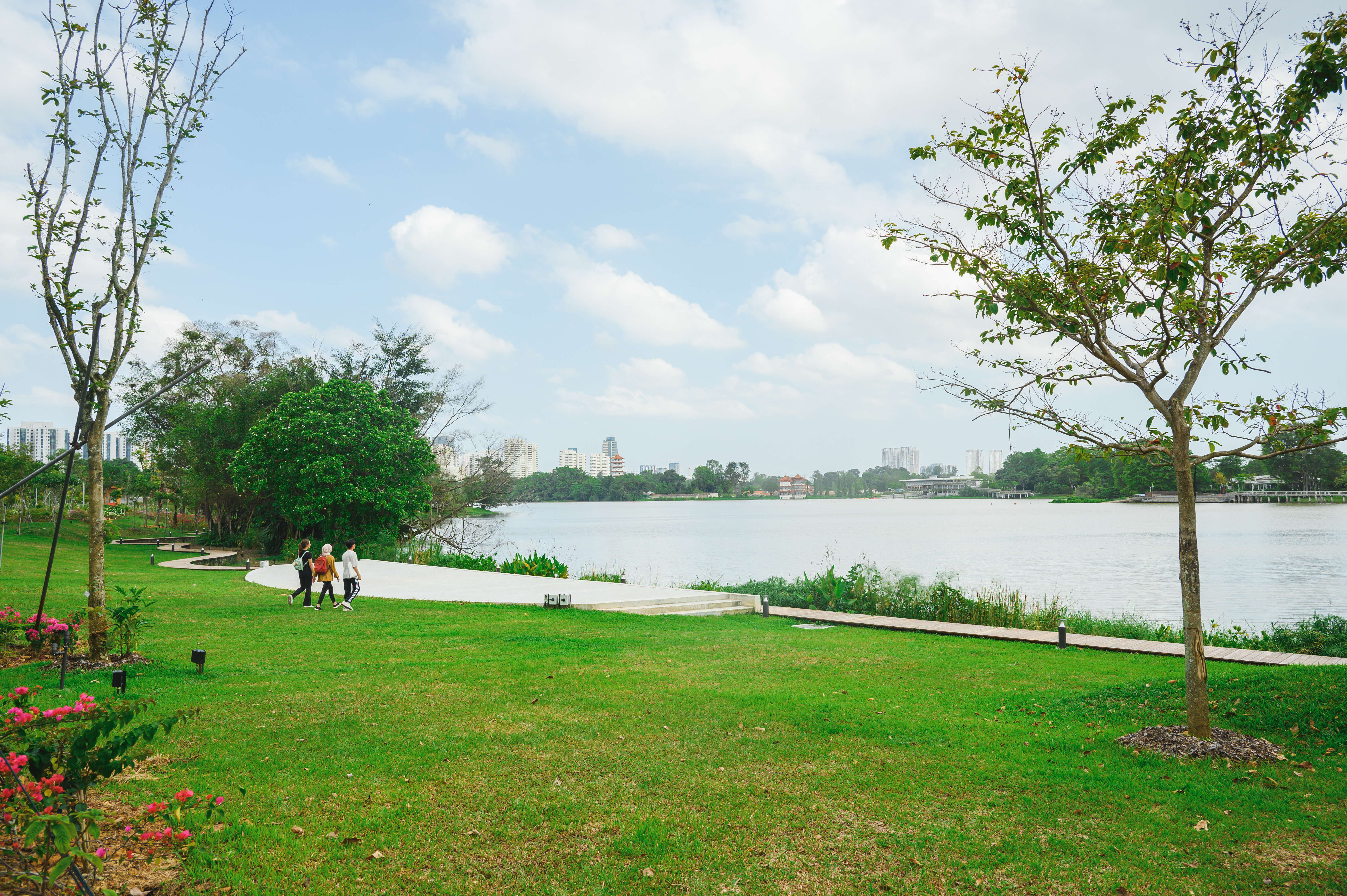 Three people walk along a path by a large lake, surrounded by trees and green grass, under a partly cloudy sky.