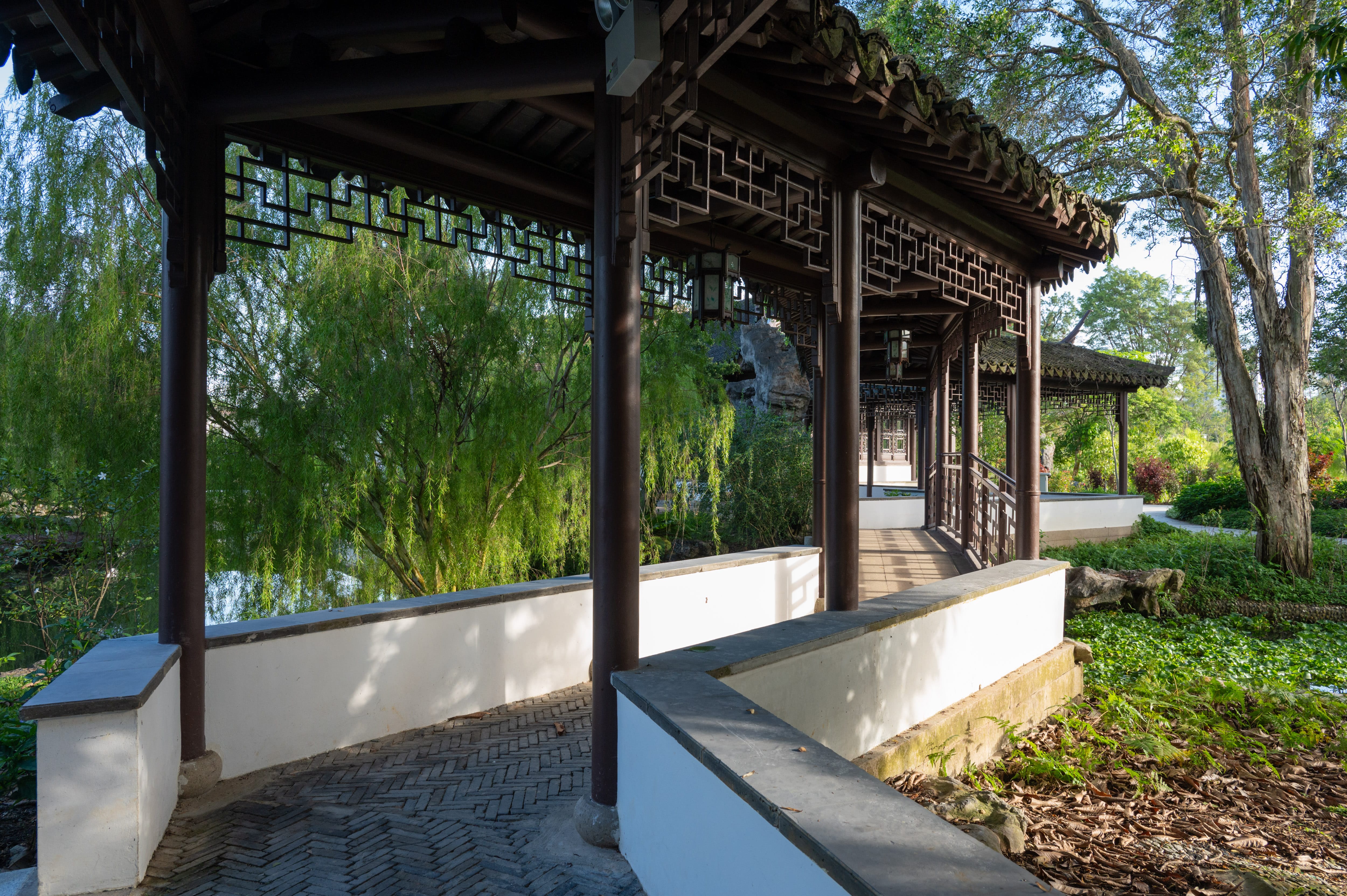 A traditional pavilion with wooden columns and latticework stands in a lush garden with trees and greenery.