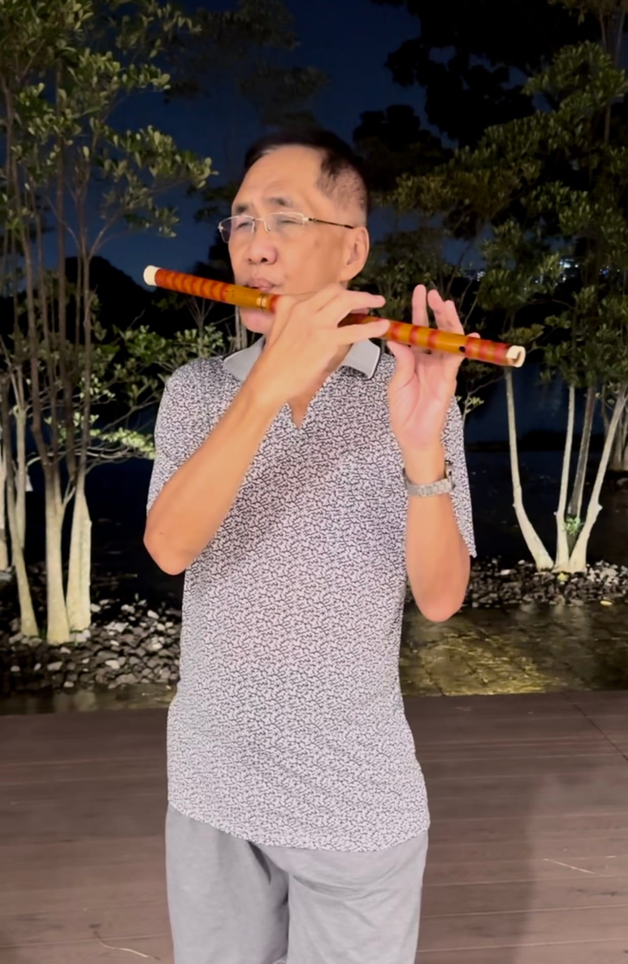 A person wearing a patterned shirt and grey pants plays a flute outdoors at night, surrounded by trees.
