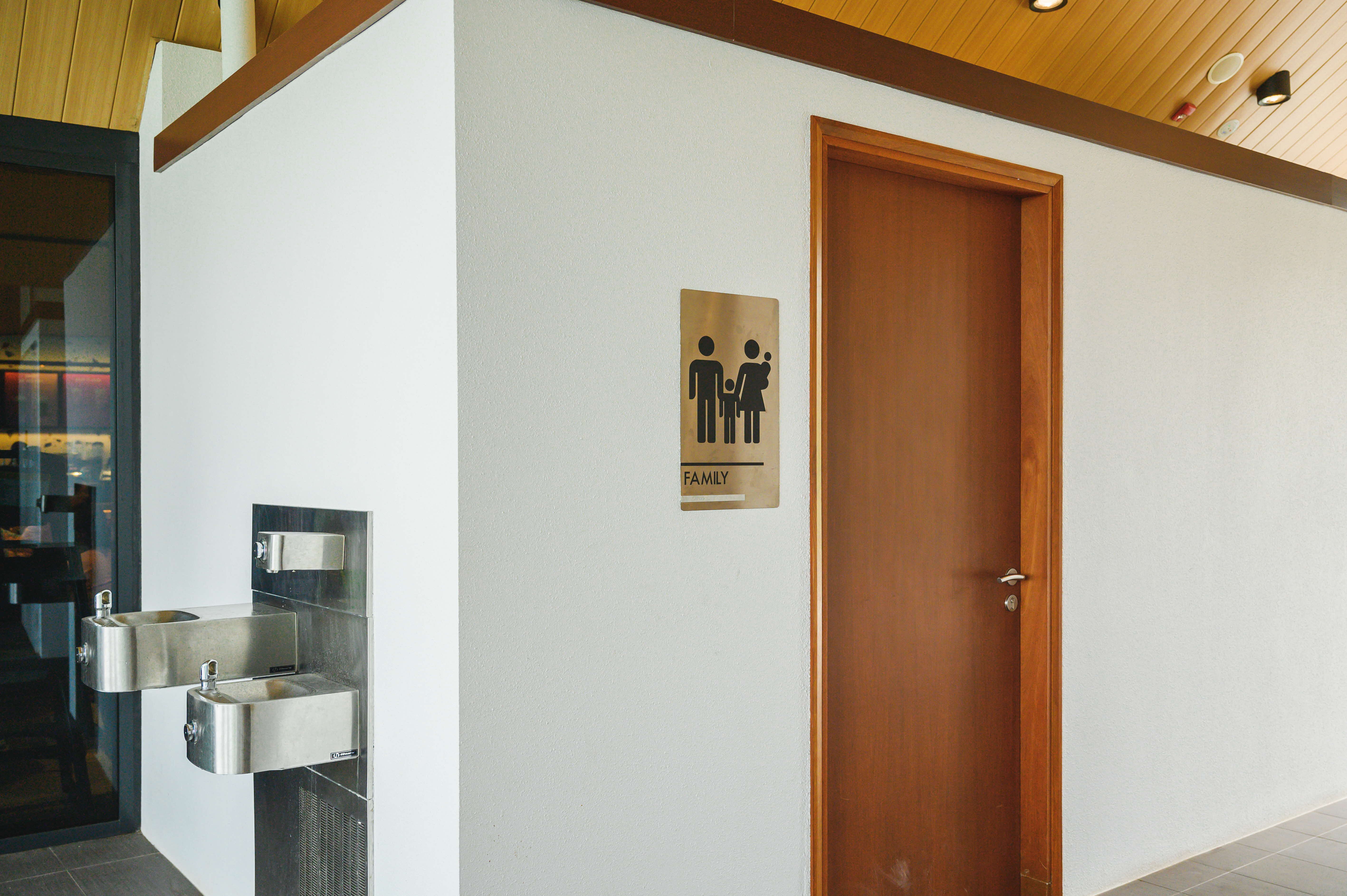 A family restroom sign is mounted on a white wall next to a wooden door, with two stainless steel water fountains attached to the wall.