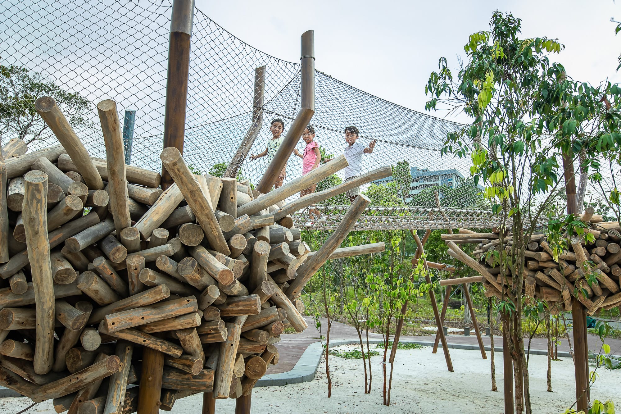 People are walking on a net bridge above wooden structures surrounded by greenery outdoors.