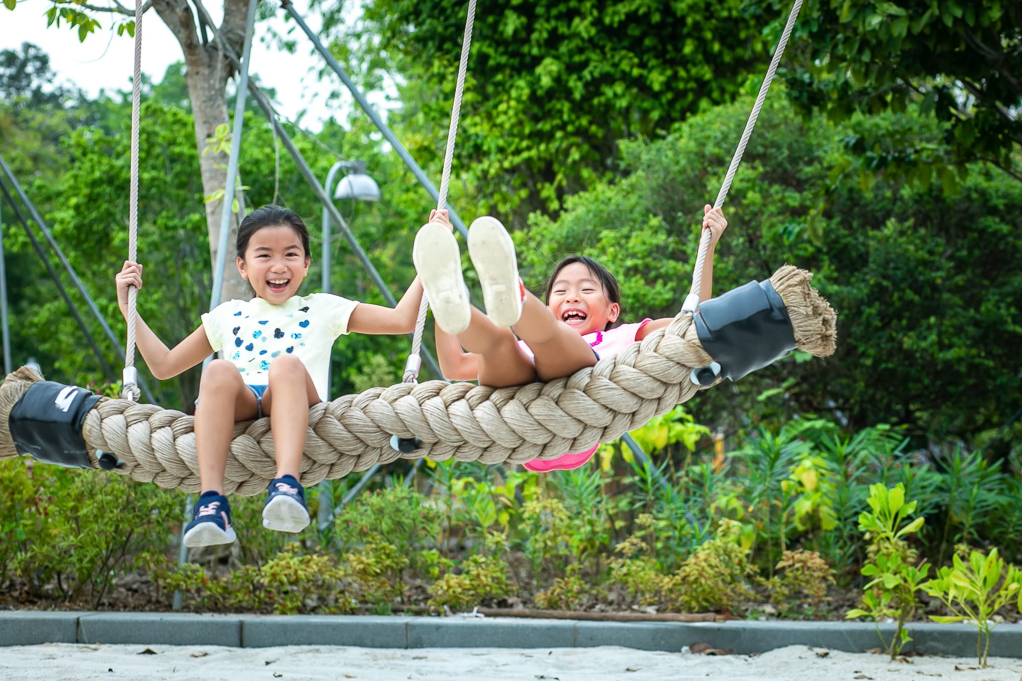 Two children sit on a large rope swing in a park surrounded by greenery and foliage.