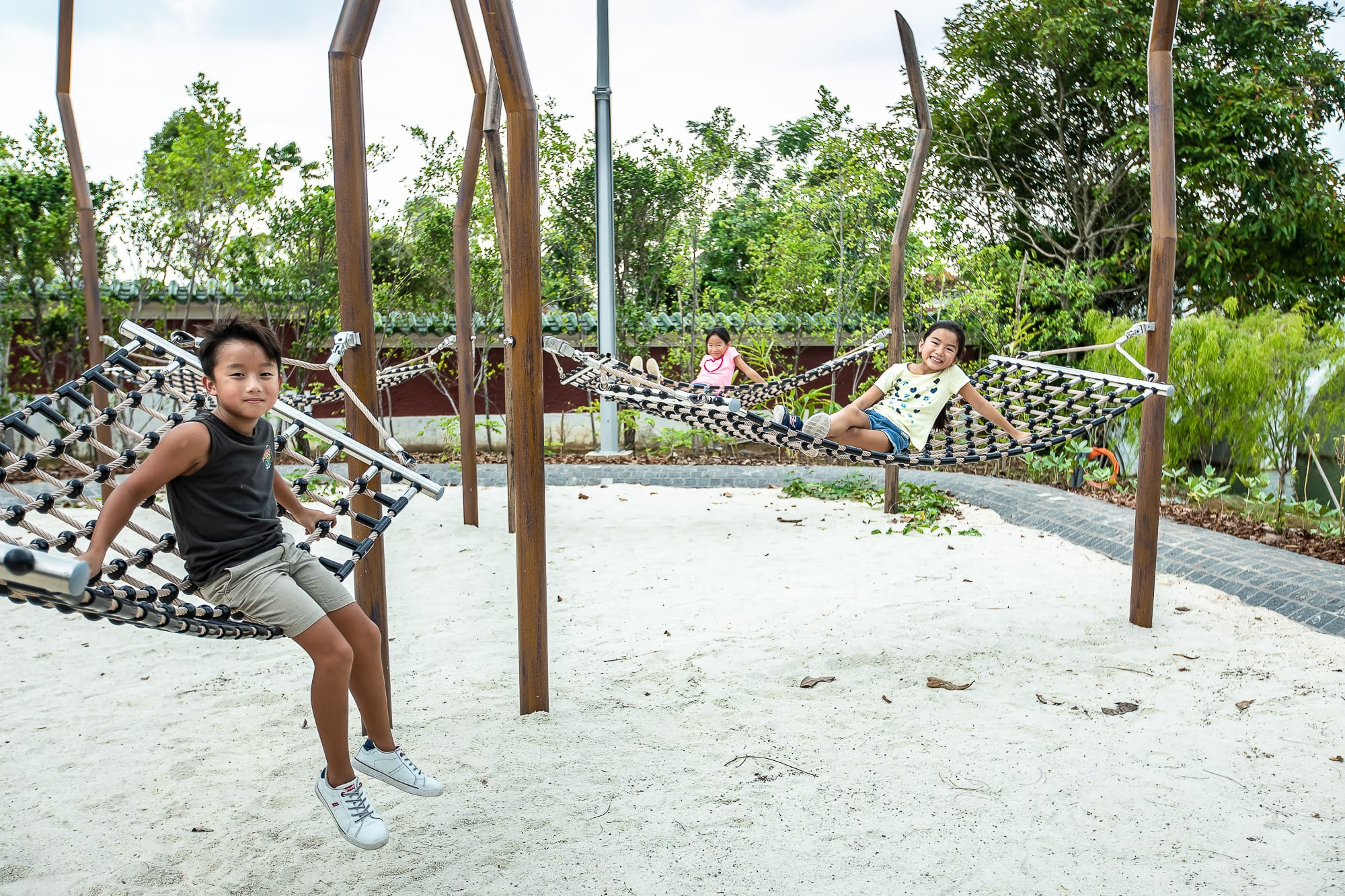 Three children sit on netted swings in a sandy outdoor area with trees and greenery in the background.